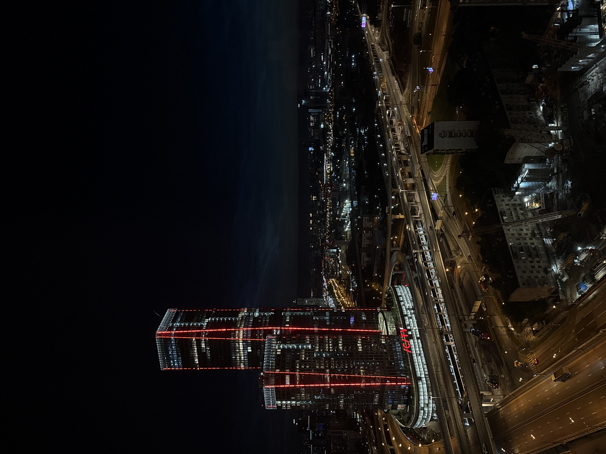 This is an aerial nighttime photograph of the Moscow City business district and surrounding infrastructure in Khimki, Russia. The prominent features are the two skyscrapers of the "City" complex, illuminated with vertical red lights, and the extensive network of highways and overpasses surrounding them. The sky is dark blue with faint traces of clouds. The skyscrapers have a modern architectural style with many windows, some of which are lit from within, indicating occupancy.
Below the skyscrapers, a multi-level highway system is visible, with cars and trucks driving on illuminated lanes. The roads are a complex network of concrete structures, overpasses, and ramps. The lighting from the vehicles and street lamps creates streaks and reflections on the wet-looking asphalt.
In the foreground and to the right of the skyscrapers, residential buildings and construction sites are visible. The residential buildings are blocky and appear to be older Soviet-era architecture. The construction sites feature cranes and piles of materials, suggesting ongoing development. The overall scene depicts a bustling urban environment at night, characterized by tall modern buildings, extensive transportation infrastructure, and ongoing construction.