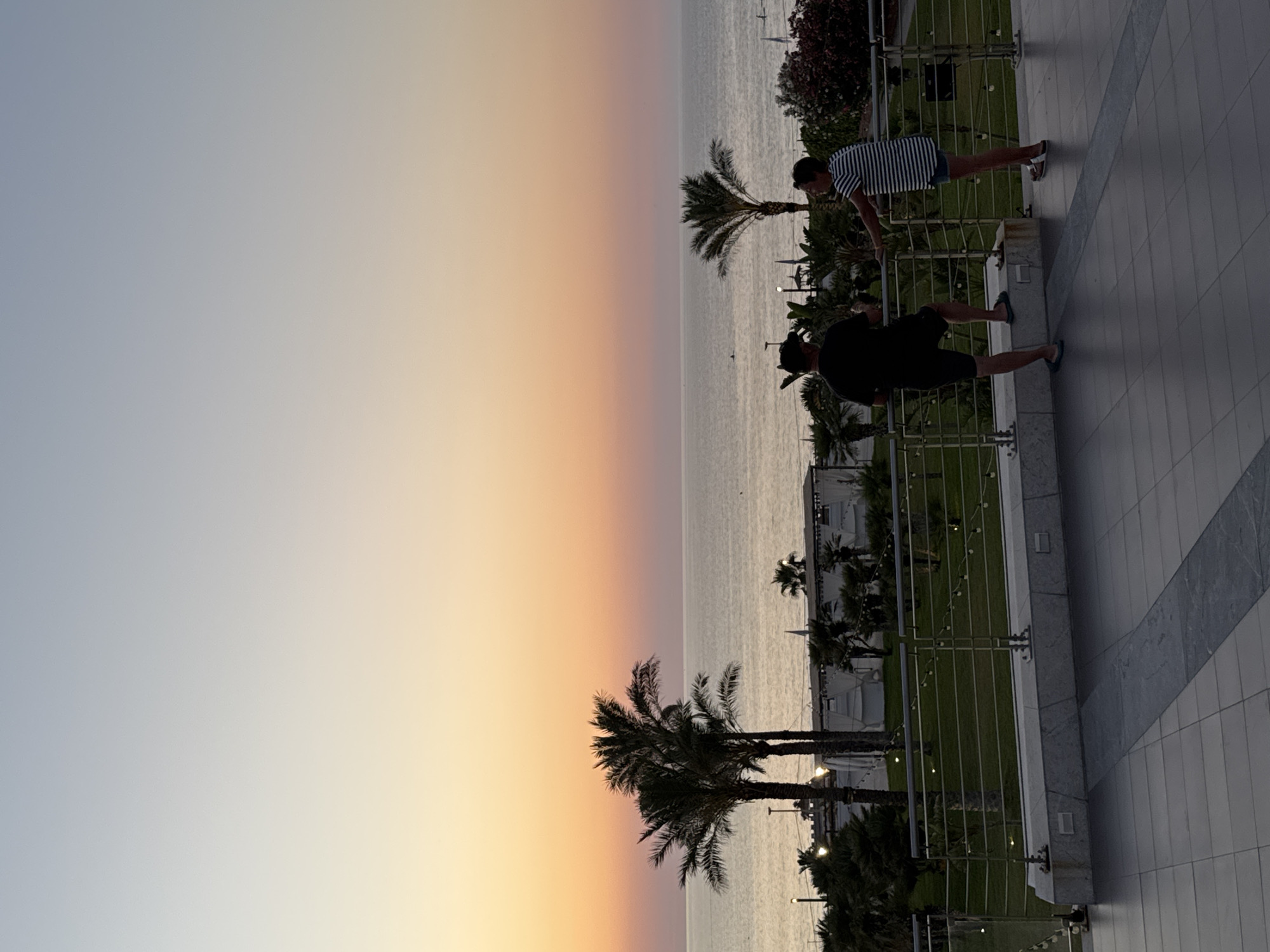 This image depicts a sunset over the sea in Isola delle Femmine, Italy. Two individuals are standing on a balcony overlooking a coastal area with palm trees and some resort-like structures. The sky displays a gradient of colors from light blue at the top to warm orange and pink hues near the horizon. The sea is calm, reflecting the colors of the sky. The balcony area has tiled flooring and a metal railing. One person is wearing a black t-shirt and shorts, while the other is in a striped t-shirt and denim shorts. They appear to be engaged in a conversation, with one leaning on the railing and the other standing nearby. String lights are visible along the balcony and in the area below.