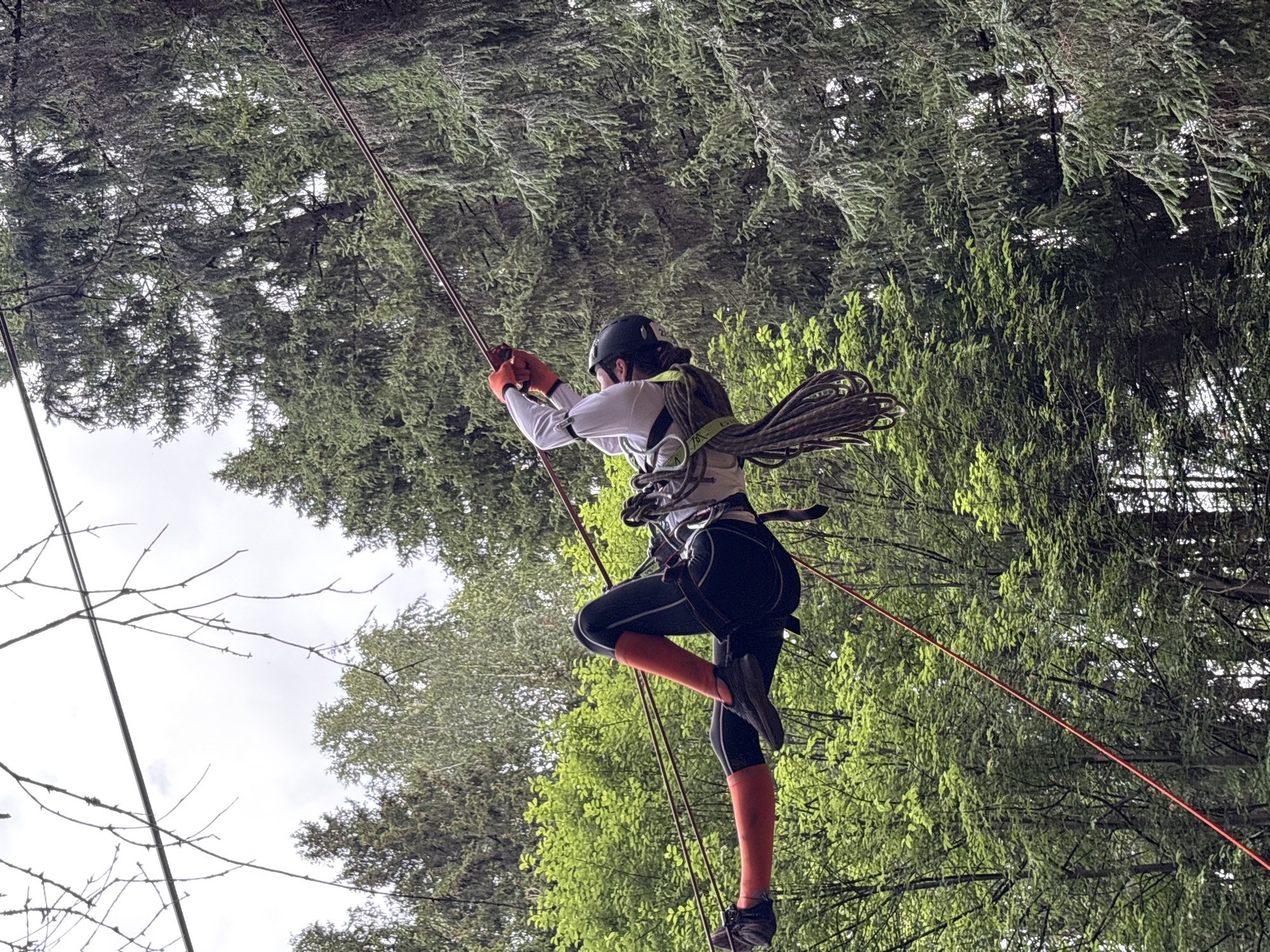 In the city of Sergino, Russia, a person is suspended mid-air, participating in an adventure activity, likely a zip line or high ropes course. The individual, wearing a white long-sleeved shirt, black pants with red accents, orange gloves, and a dark helmet, is secured by ropes and a harness. They are positioned horizontally, with their legs extended. A thick coil of rope hangs from their harness, and a thinner red rope is visible diagonally across the frame. The background consists of lush green trees, suggesting an outdoor, natural setting within the city. The sky above is overcast.