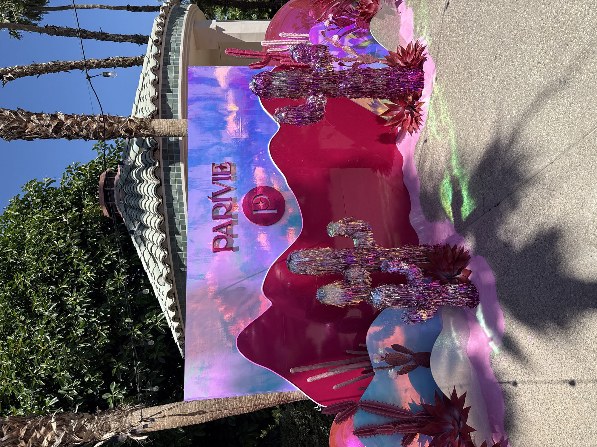 The image shows a festive, oversized display in La Quinta, California. The display is primarily pink, blue, and purple, and is decorated with metallic-fringe cactus shapes. The name "PARVIE" is printed vertically on a portion of the display with a gradient sky effect. The backdrop resembles a stylized desert landscape. The display is set up outside near a building with a tiled roof and palm trees, with sunlight casting shadows on the adjacent paved area.