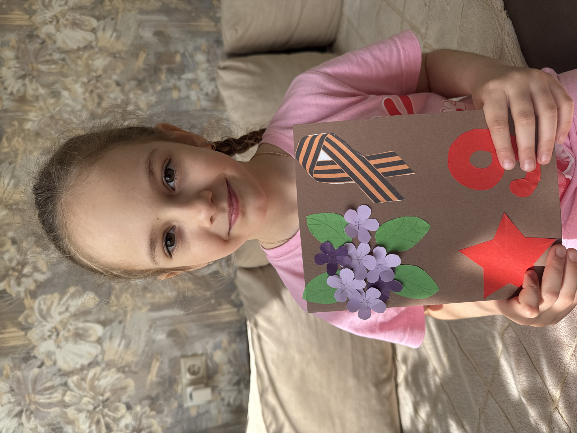 A young girl with braided hair is sitting on a beige couch, holding a brown card decorated with paper cutouts. The cutouts include a black and orange ribbon, the number "9" in red, purple flowers with green leaves, and a red star. The girl is wearing a pink t-shirt. The background includes a wall with a floral pattern. The location is identified as the city of Раздолье, Russia.