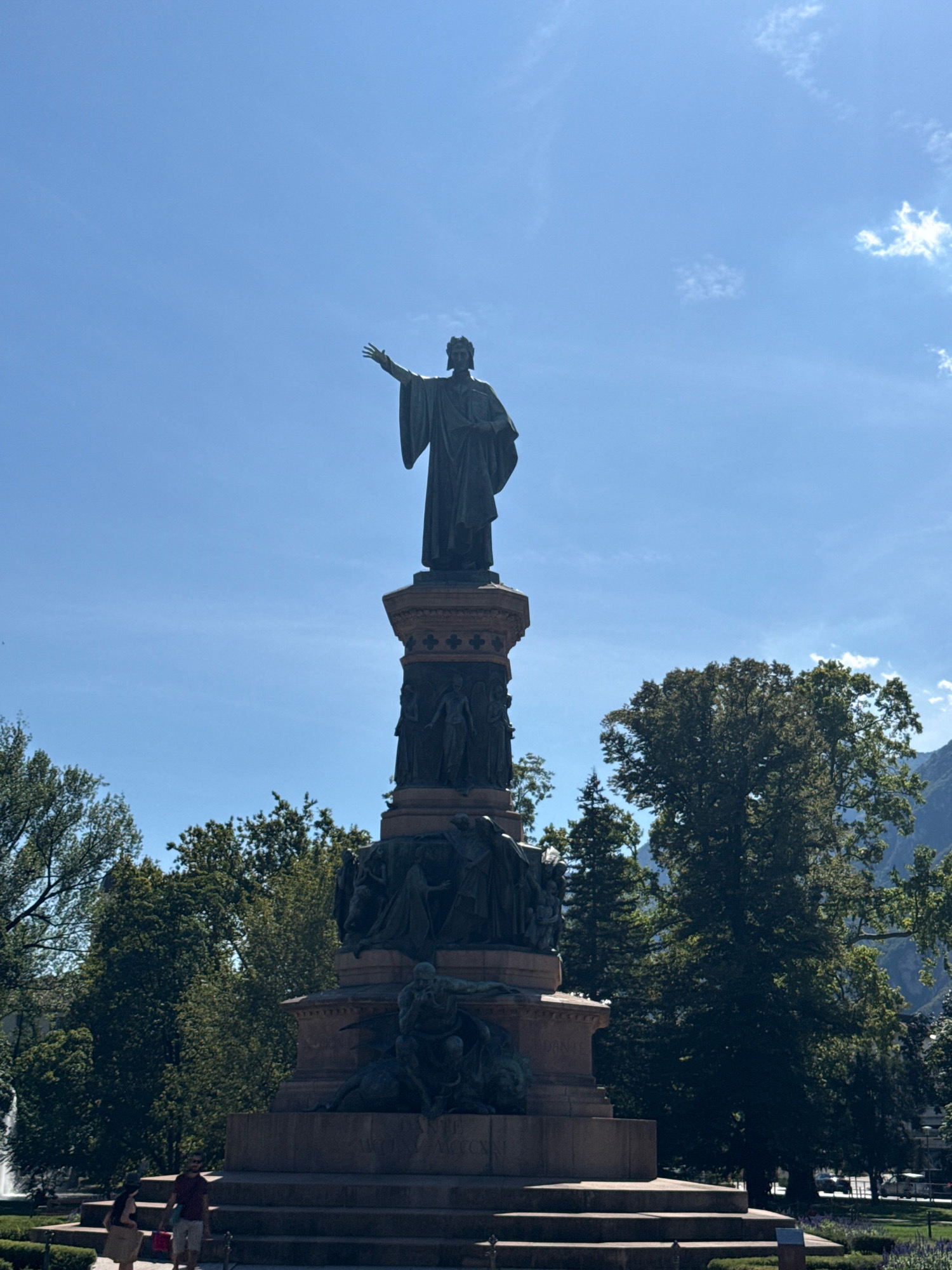 The image shows a large bronze monument in Trento, Italy, under a bright blue sky with scattered clouds. The monument features a central figure at the top, a man with a crown, wearing a robe and pointing with his left hand. Below this figure is a decorated pedestal with multiple relief sculptures depicting human figures. The base of the monument is a large stone structure with tiered steps.
In the foreground, on the steps of the monument, two individuals are visible. A woman on the left is wearing a light-colored dress, and a man on the right is wearing a maroon t-shirt and white shorts, carrying shopping bags.
Surrounding the monument are lush green trees, with some taller trees providing a backdrop. In the far left background, a fountain is partially visible. In the far right background, mountains can be seen, and a street with some vehicles is also present. The overall scene suggests a sunny day in a public park or square in Trento.