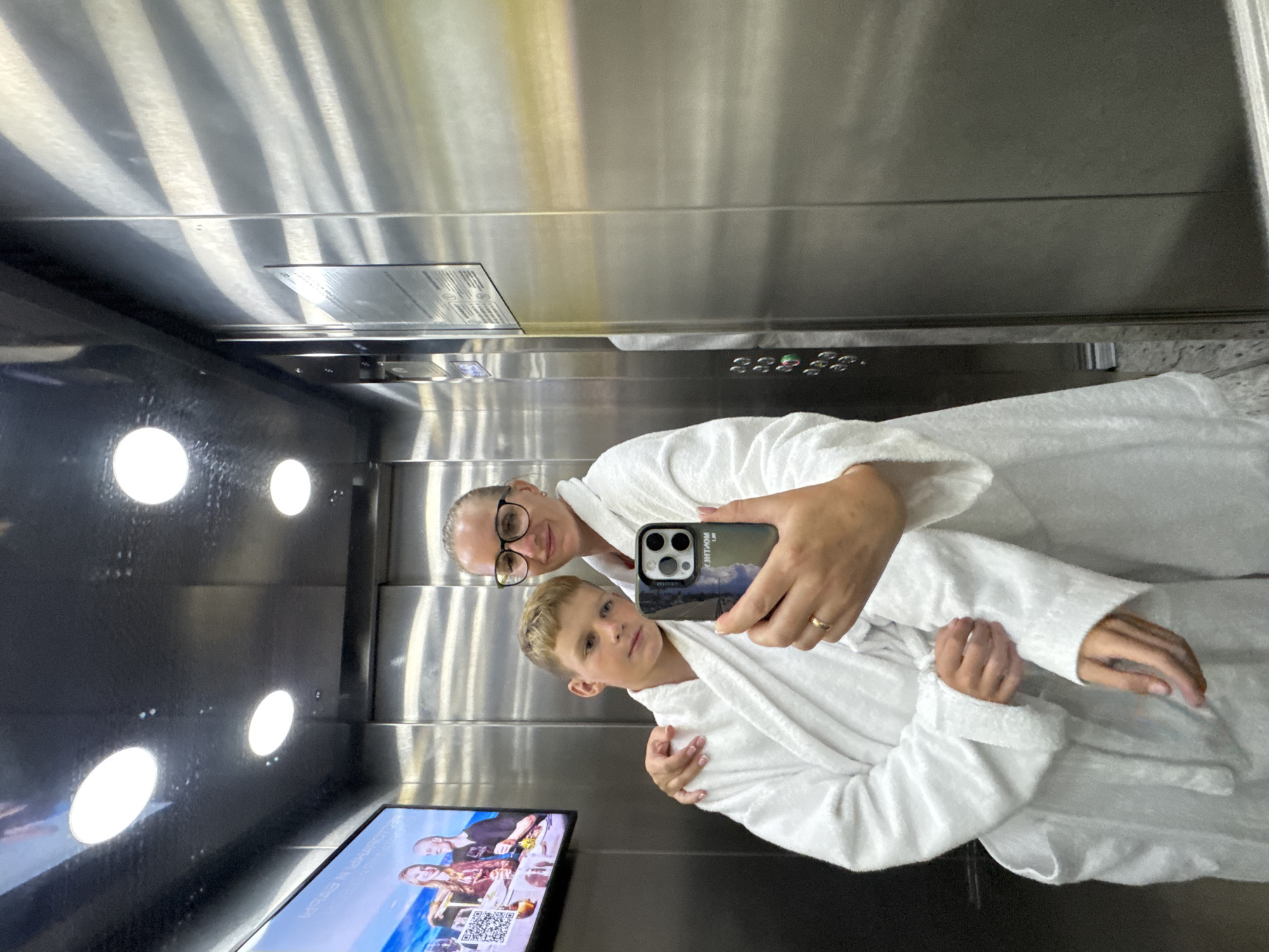 A woman and a boy, both wearing white bathrobes, are taking a selfie in an elevator. The woman is wearing glasses and has her arm around the boy. She is holding a smartphone, angled to capture their reflection in the elevator's mirrored walls. The boy is looking towards the camera with a neutral expression. The elevator interior is made of stainless steel, with visible control buttons on one wall and a screen displaying an advertisement on the opposite wall. The lighting is provided by overhead circular fixtures, casting reflections on the metallic surfaces. This scene is in the city of Vityazevo, Russia, likely within a hotel or resort.