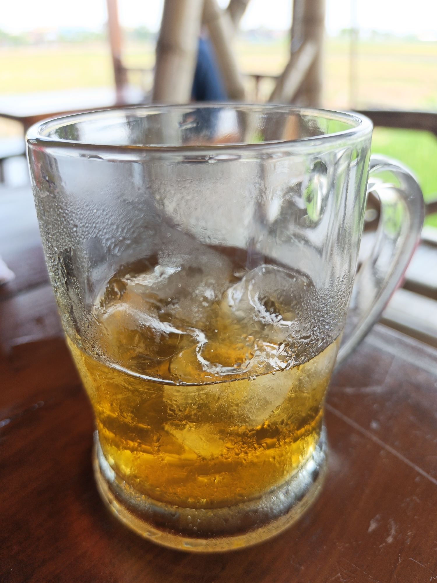 An eye-level, close-up shot shows a clear glass mug filled with iced tea and ice cubes, sitting on a wooden table. The mug's exterior is covered in condensation. In the background, out of focus, are bamboo structures and green foliage, suggesting an outdoor setting possibly in Sekarsuli, Indonesia, given the environmental cues. A blurred figure of a person is visible in the far background.