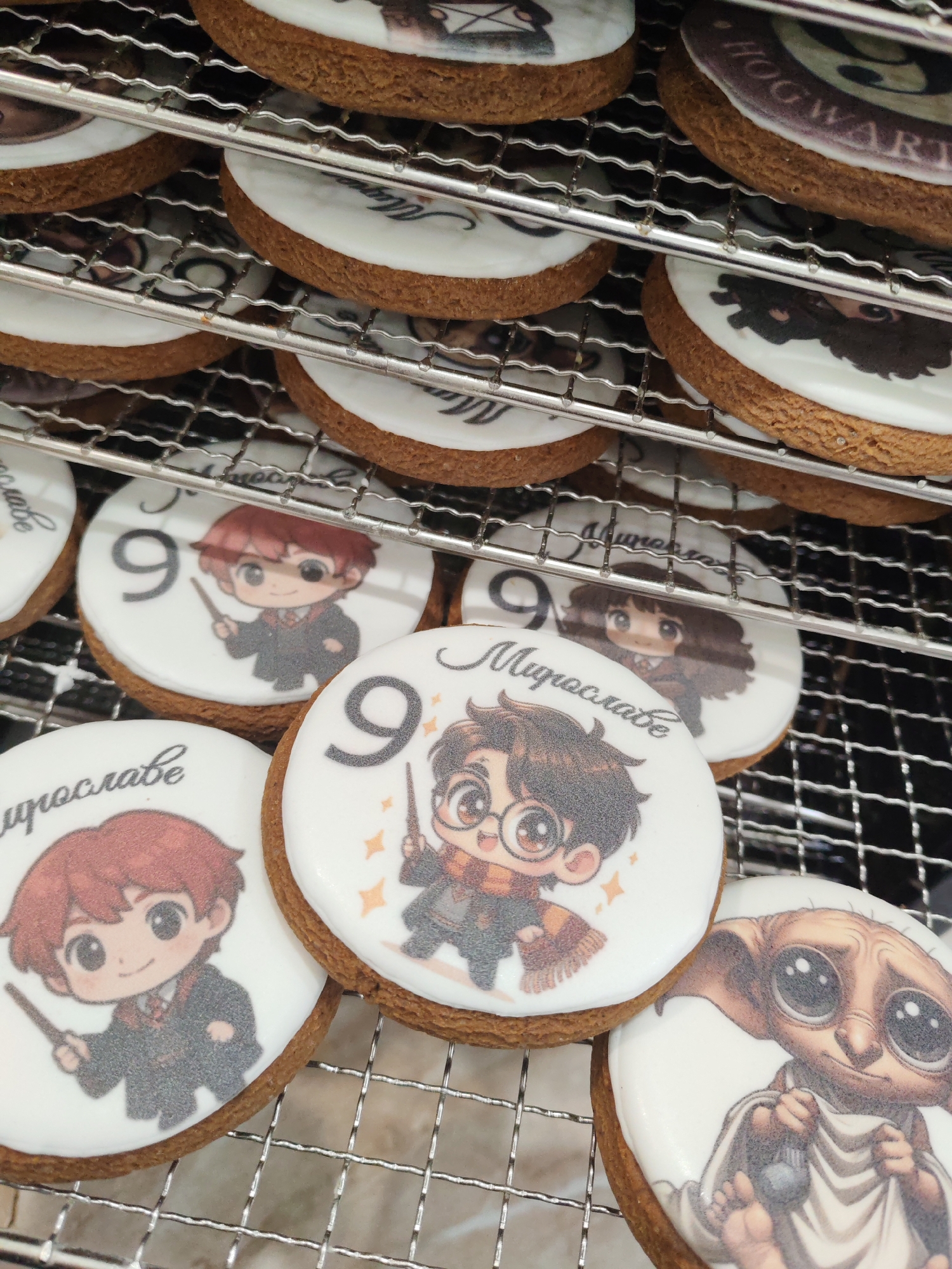 The image shows a set of round cookies decorated with images related to the Harry Potter franchise, placed on a metal cooling rack. The cookies feature cartoon illustrations of characters such as Harry Potter, Ron Weasley, Hermione Granger, and Dobby. Some of the cookies also include the number "9" and the name "Myroslave" written in Cyrillic. The cookies have a light-colored frosting on top, serving as a background for the images, and a dark brown, textured cookie base. The cooling rack has multiple tiers and a wire mesh pattern.