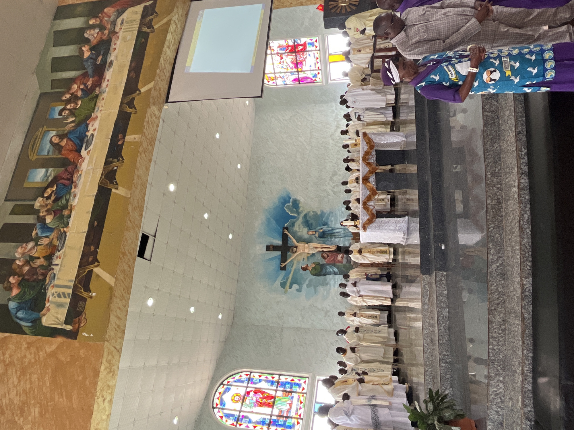 The image depicts the interior of a church, likely in Vom, Nigeria, during a religious service. Dominating the background is a large mural of "The Last Supper" on the upper wall. Below this, a prominent crucifix with Jesus Christ is displayed, flanked by painted figures. Stained-glass windows, one featuring a religious scene, are visible on the left and right walls. A projection screen hangs from the ceiling. In the foreground, a group of individuals, predominantly men in white and cream ceremonial robes, stand behind an altar. Two women in traditional attire are positioned in front of the altar. The church features tiered steps leading to the altar and a polished floor.