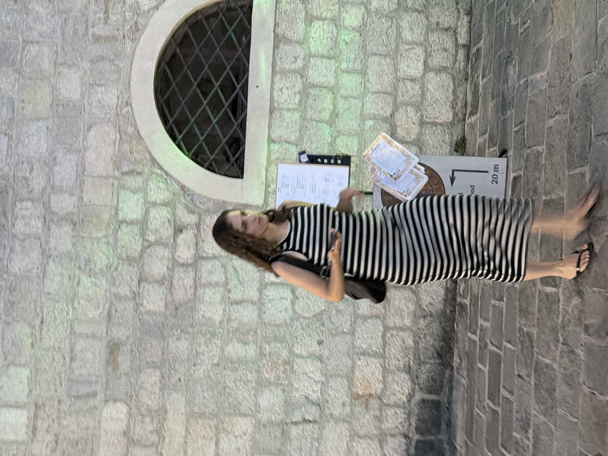 A young woman with curly brown hair walks on a stone-paved sidewalk in front of a stone wall. She is wearing a sleeveless black and white striped maxi dress and black flip-flops. She carries a black backpack and holds a smartphone in her right hand, and a stack of menus in her left hand. To her right, there is a sign with text and an arrow pointing left, indicating a distance of "20 m." Above the sign and to the right, there is a semicircular arched window with a dark metal grate. The stone wall is constructed from large, rough-cut blocks, and a soft green light illuminates portions of it. The scene appears to be outdoors in a city, possibly in Budva, Montenegro, given the architectural style. The woman is in motion, with her legs blurred, suggesting she is walking.