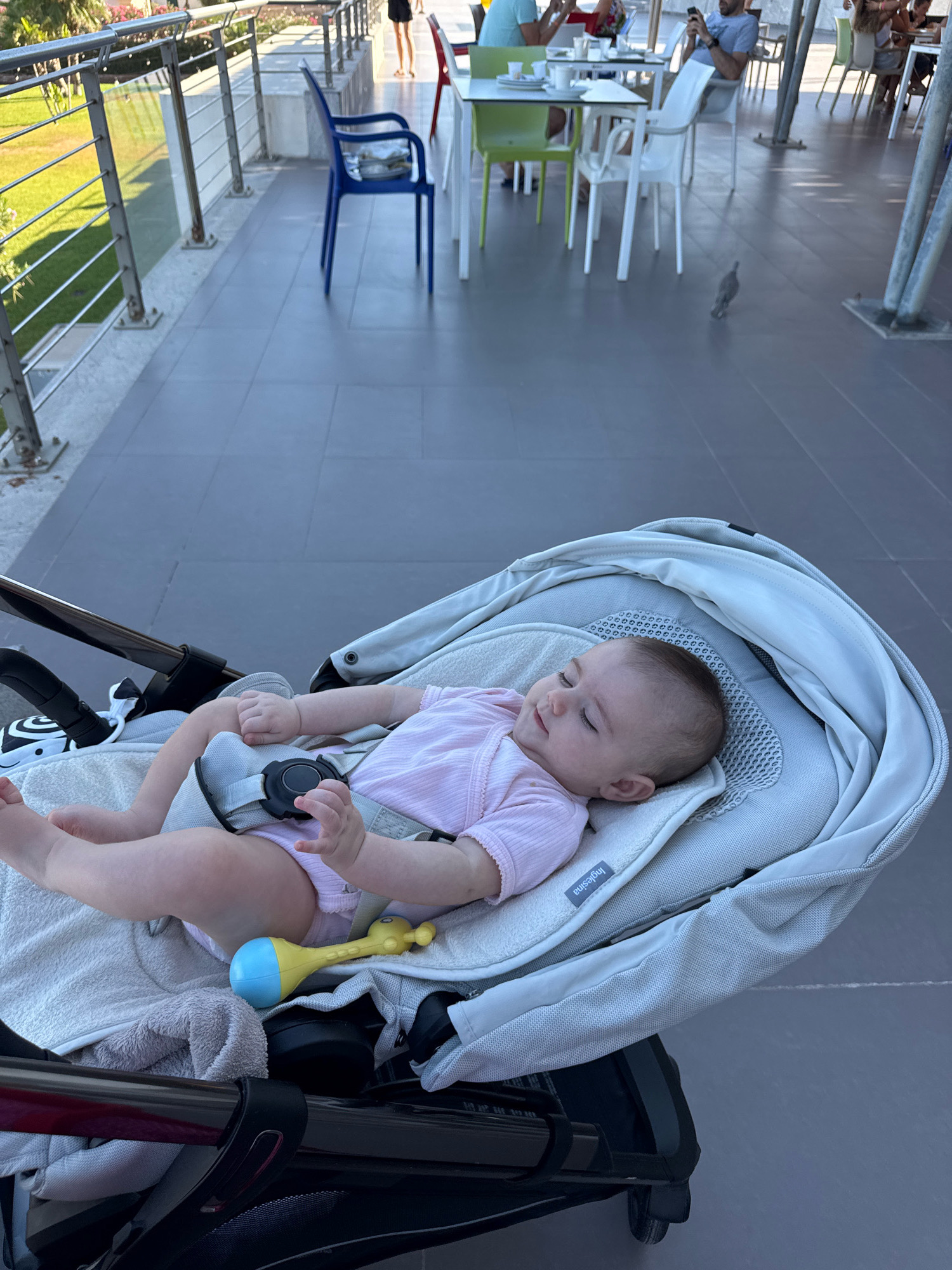 An outdoor paved area features a baby lying in a stroller. The baby is wearing a pink ribbed outfit and appears to be sleeping. A yellow and blue rattle is placed near the baby. The stroller is light gray with black accents. In the background, several people are seated at tables in what appears to be an outdoor seating area of a restaurant or cafe. Green and white chairs are visible around the tables. A blue plastic chair is situated on the paved area. A bird is visible on the ground in the mid-ground. A railing with horizontal bars runs along the left side of the image, overlooking a grassy area and trees. The location is Isola delle Femmine, Italy.