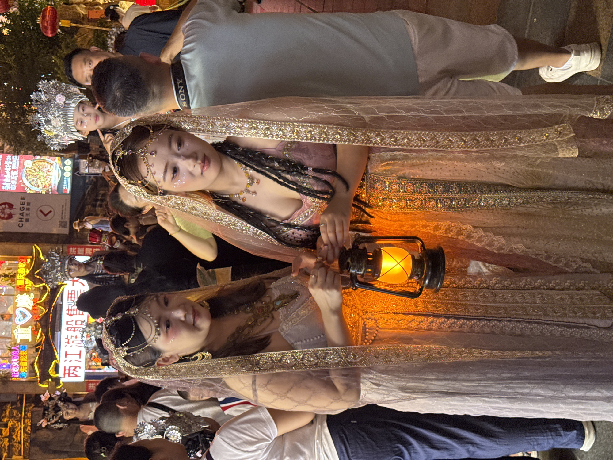 Two women dressed in elaborate traditional Chinese costumes, adorned with veils and intricate jewelry, stand in a crowded outdoor night market. The woman on the left wears a light-colored dress and holds an antique lantern that emits a warm orange glow. The woman on the right is dressed in a pink garment with a similar veil and jewelry. They are surrounded by a bustling crowd of people, some of whom are also in costume, suggesting a cultural festival or event. In the background, illuminated signs with Chinese characters indicate the location is in Yuzhong District, China, a vibrant urban area known for its nightlife and commercial activity. The overall atmosphere is festive and visually rich, with neon lights and traditional decorations contributing to the scene.