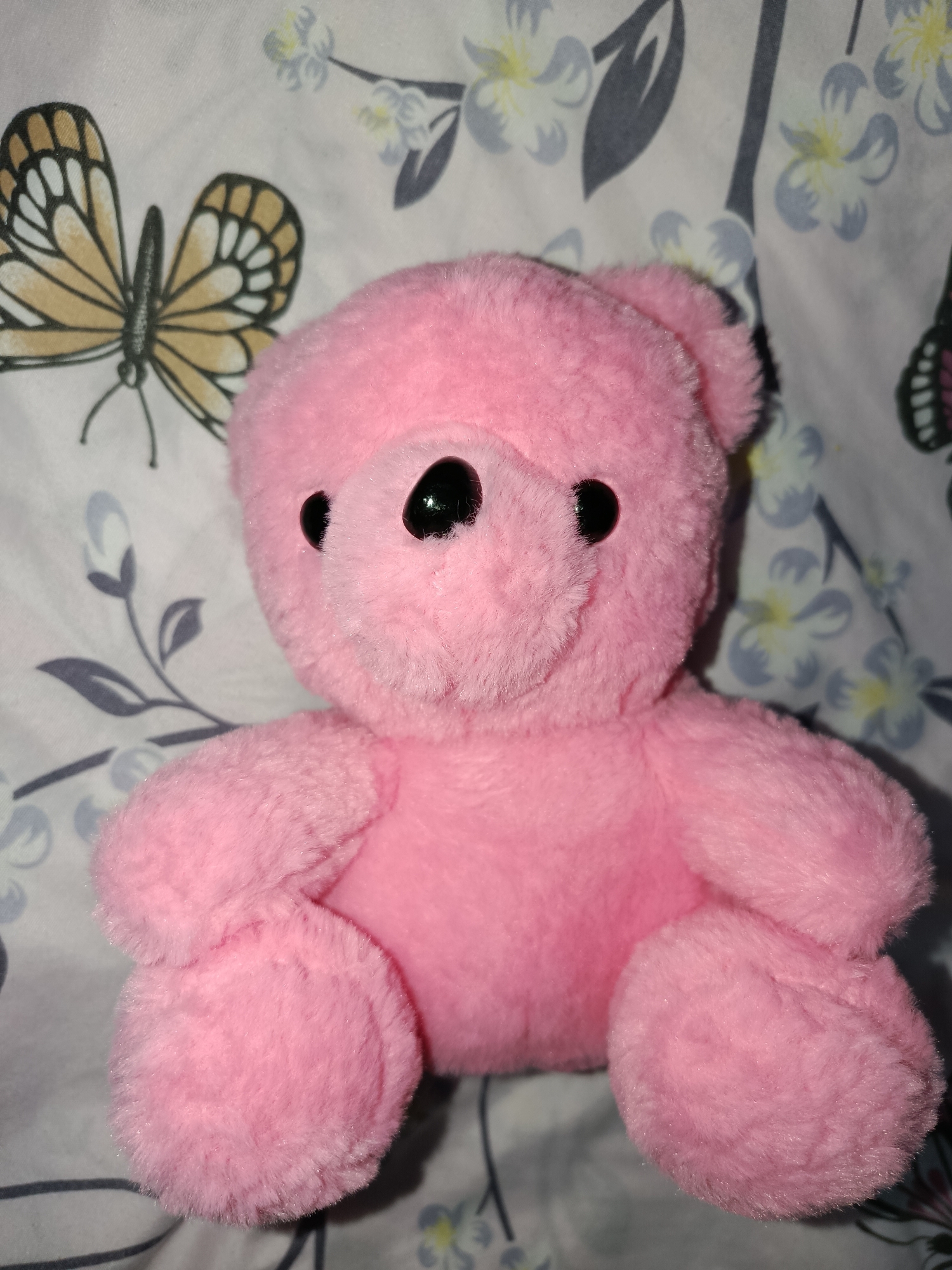 The image features a close-up shot of a small, fluffy pink teddy bear. The bear is positioned centrally, facing the viewer, with its paws resting on its lap. It has black plastic eyes and a small black nose. The background is a light-colored fabric with a repeating pattern of stylized flowers and butterflies. The flowers are depicted in shades of gray, blue, and yellow, with some appearing as buds and others in full bloom. The butterflies are a warm beige color with black outlines and intricate wing patterns. The overall impression is that of a child's toy placed against decorative fabric, possibly a blanket or curtains. There is no visible text in the image. The lighting suggests an indoor setting, likely in daylight, but no specific time of day or weather conditions can be inferred. The location context provided is Tashkent, Uzbekistan, but there are no visual cues within the image to confirm this specific location. The emotion conveyed is one of innocence and gentleness, typical of a teddy bear.