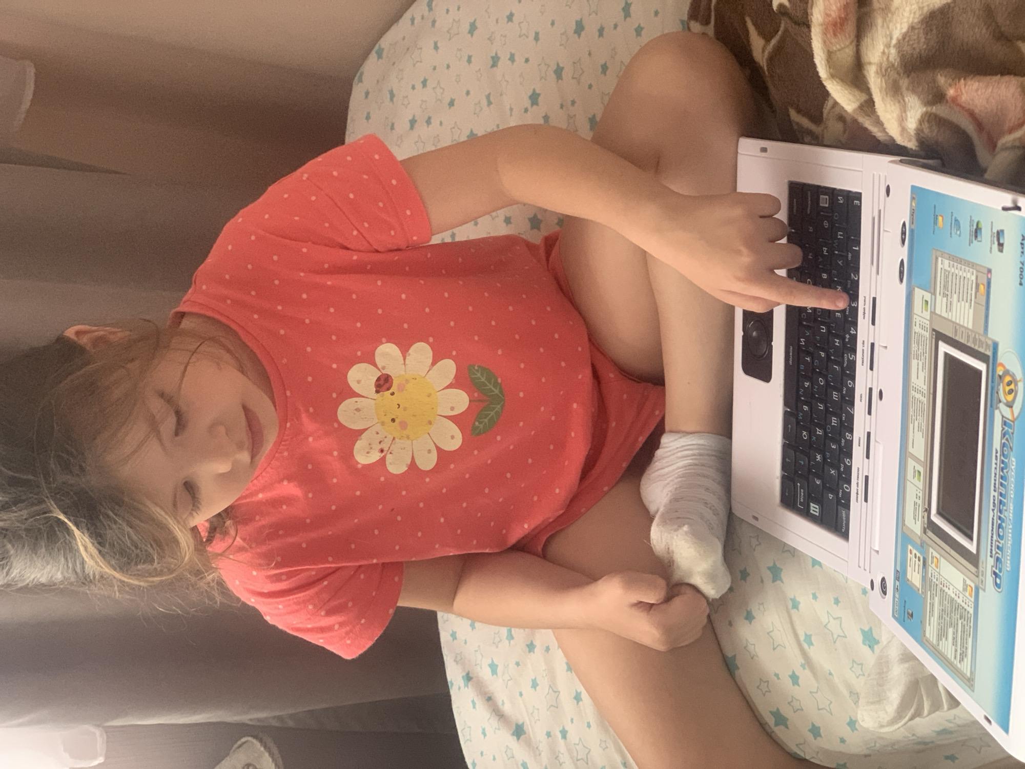 A young girl is lying on a bed, playing with a toy laptop. She is wearing a pink t-shirt with white polka dots and a flower design. The laptop is white and has a keyboard with Cyrillic letters. The screen displays what appears to be a children's educational program. The girl is looking down at the laptop with a relaxed expression. The scene is indoors, with a blanket draped over the bed and a pillow with a star pattern. The lighting suggests it could be daytime or early evening. The girl's position and the presence of the toy laptop indicate she is engaged in play or learning. The visible text on the laptop includes "KOMUGP" and "АВТСКИЙ ОБУЧ", which suggests it's a Russian educational toy laptop.