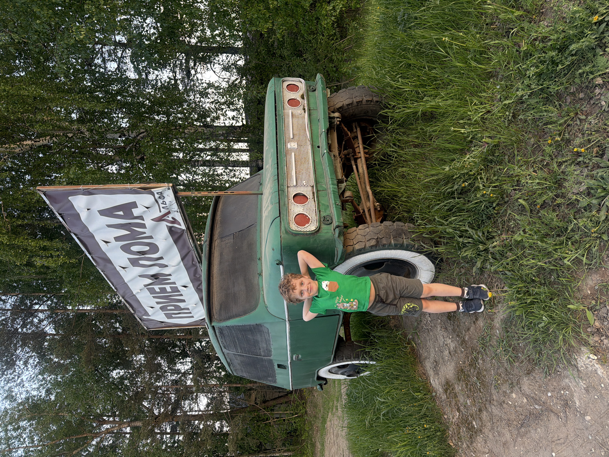 A medium shot presents a boy lying down next to an overturned, green, vintage vehicle, likely a truck, in a grassy, wooded area. The boy has curly blond hair and is wearing a green t-shirt with a cartoon-like graphic, khaki shorts with a design on the knee, and black sneakers with white soles. He is lying on his back with his hands behind his head and appears to be looking at the camera with a slight smile.
The truck is upside down and has large, knobby tires. A white spare tire rests next to the boy. The truck's rear end is facing the camera, revealing the tail lights and a chrome grill. A large banner with white Cyrillic text on a dark background is attached to the truck.
The surrounding environment is a dirt path and dense green foliage, suggesting the photo was taken in a forest or park. Based on prior knowledge of the phrase on the banner, the location is likely the city of Chaikovsky, Russia. The lighting suggests it is daytime.