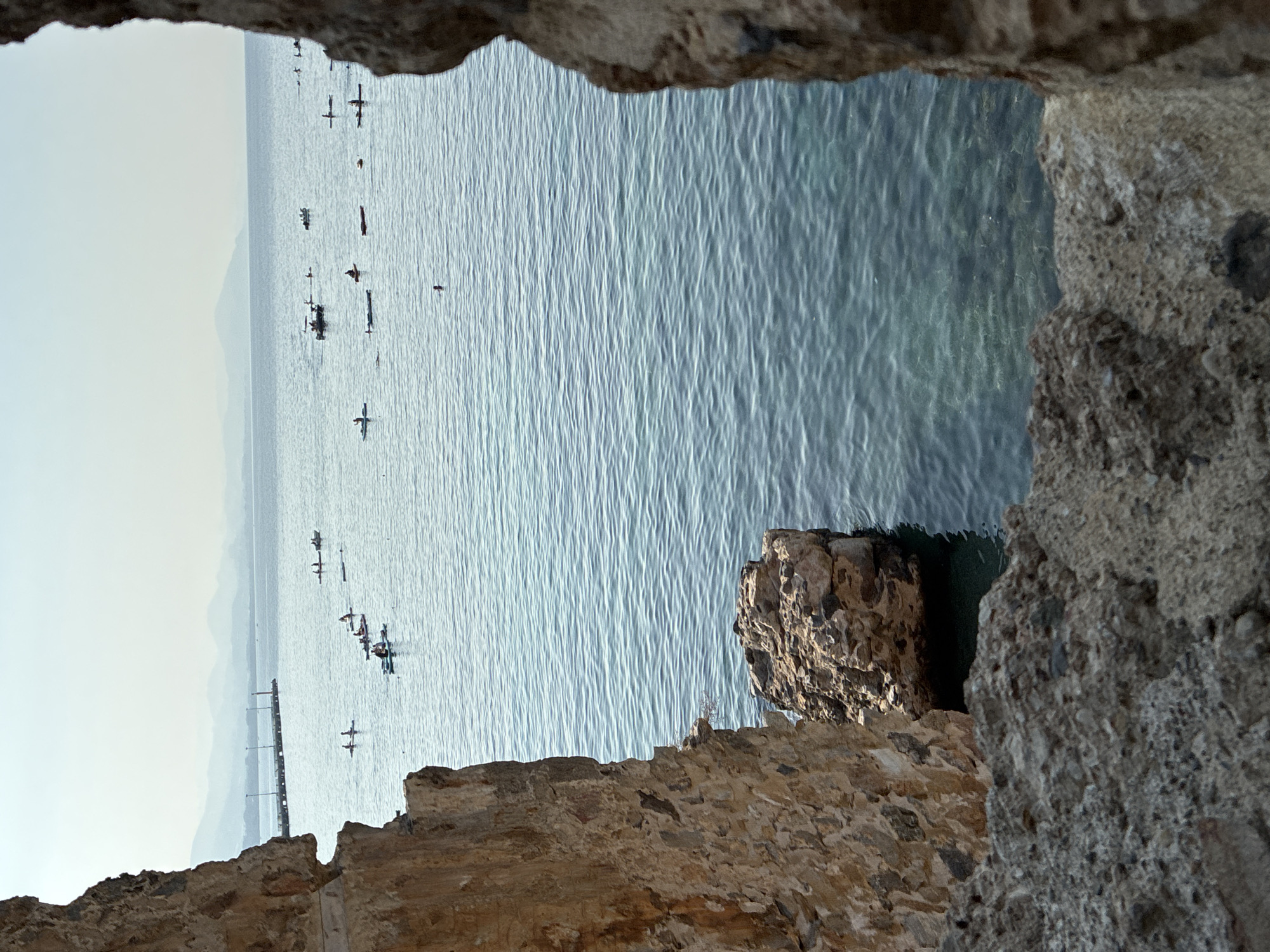 The image shows a view of the Mediterranean Sea from within an ancient stone structure, likely part of the Alanya Castle walls, in Alanya, Turkey. The sea is calm with gentle ripples, and the water is a clear, greenish-blue, allowing visibility of the seabed in some areas.
Numerous people are engaged in various water activities. Several individuals are on stand-up paddleboards, while others are in kayaks or small boats. Some are actively paddling, while others appear to be resting or observing. One person is visible swimming in the distance.
In the background, a pier extends into the sea with lights and structures on it. Beyond the pier, hazy mountains form the horizon, with a pale sky above. The foreground is dominated by rough, textured stone walls and rocks, framing the view of the sea. The stone appears ancient, with varying shades of brown and grey. A large, jagged rock formation juts out into the water on the left side of the frame, near the base of the stone structure. The scene suggests a recreational day at the sea, viewed from a historical vantage point.