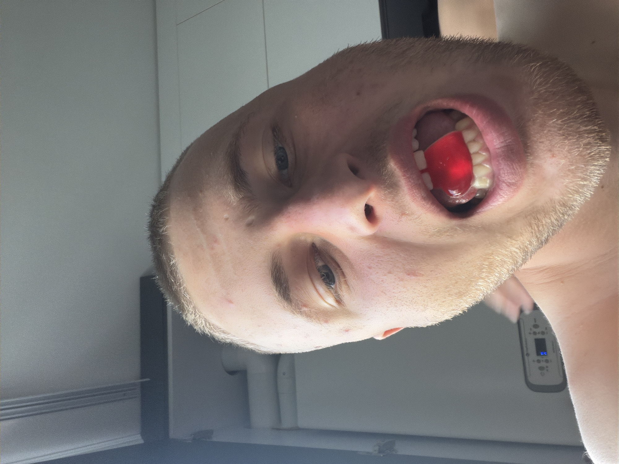 A young man is captured in a close-up, low-angle selfie, with a red gummy candy held between his teeth and tongue. He has short, light brown hair and fair skin with visible blemishes. His mouth is open, revealing his teeth and the vibrant red candy. The background appears to be an indoor setting with a neutral-colored wall and a hint of what might be a window frame. There is no discernible indication of the city of Strășeni, Moldova within the image.
