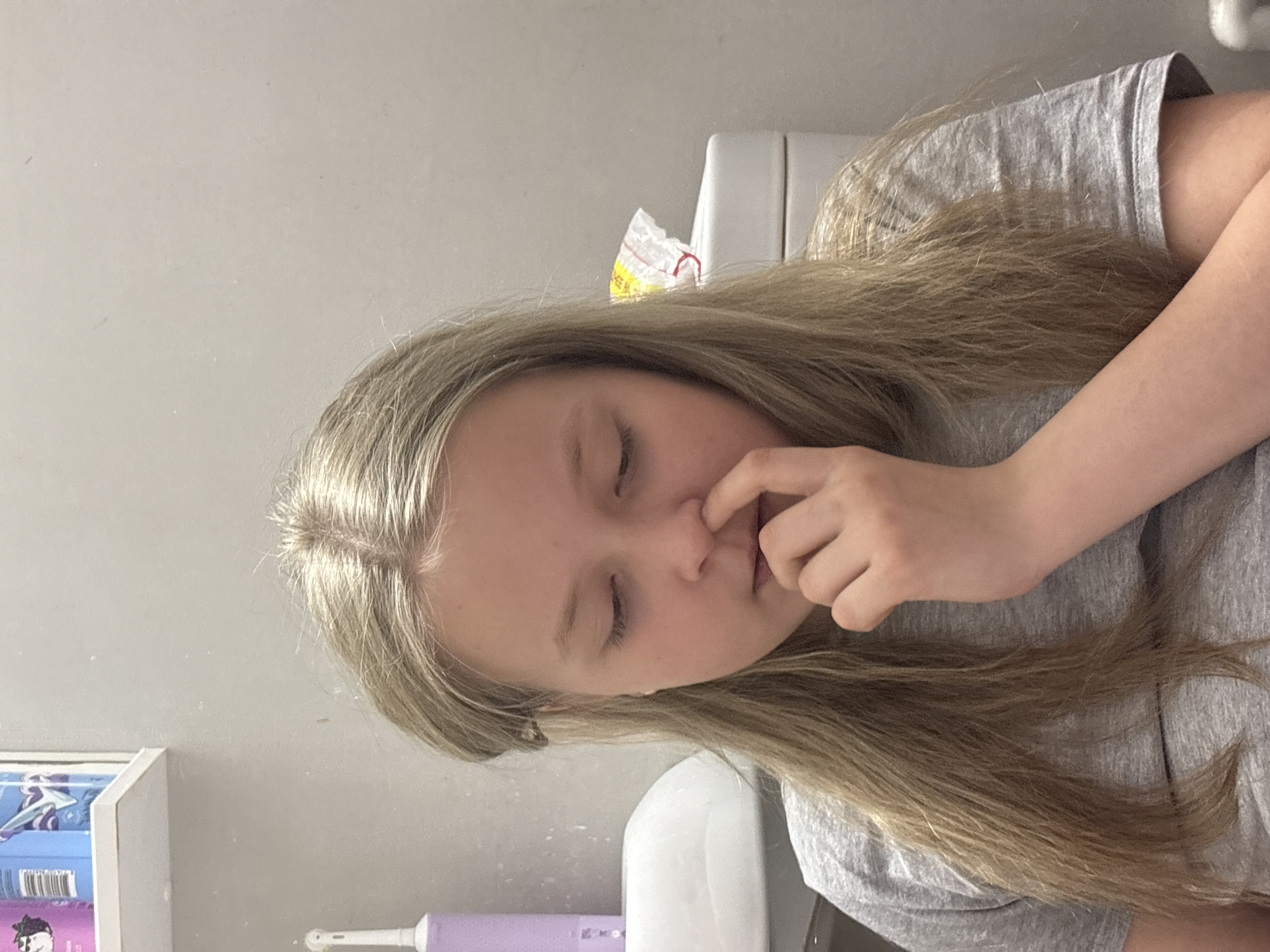 A young person with fair skin and light brown hair is picking their nose. They are wearing a gray t-shirt. The background includes a white wall, a toilet, and a shelf with items on it. There is no evidence to suggest that the image was taken in Izhevsk, Russia.
