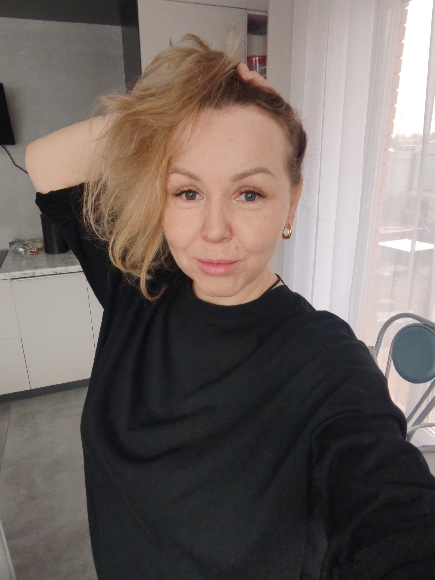 A fair-skinned woman with blonde hair and freckles is taking a selfie indoors. She is wearing a black long-sleeved shirt and gold hoop earrings. Her left hand is raised, touching her hair. The background includes kitchen cabinets, a window with sheer curtains, and a metal chair. The location is suggested to be in the city of Chaikovsky, Russia.