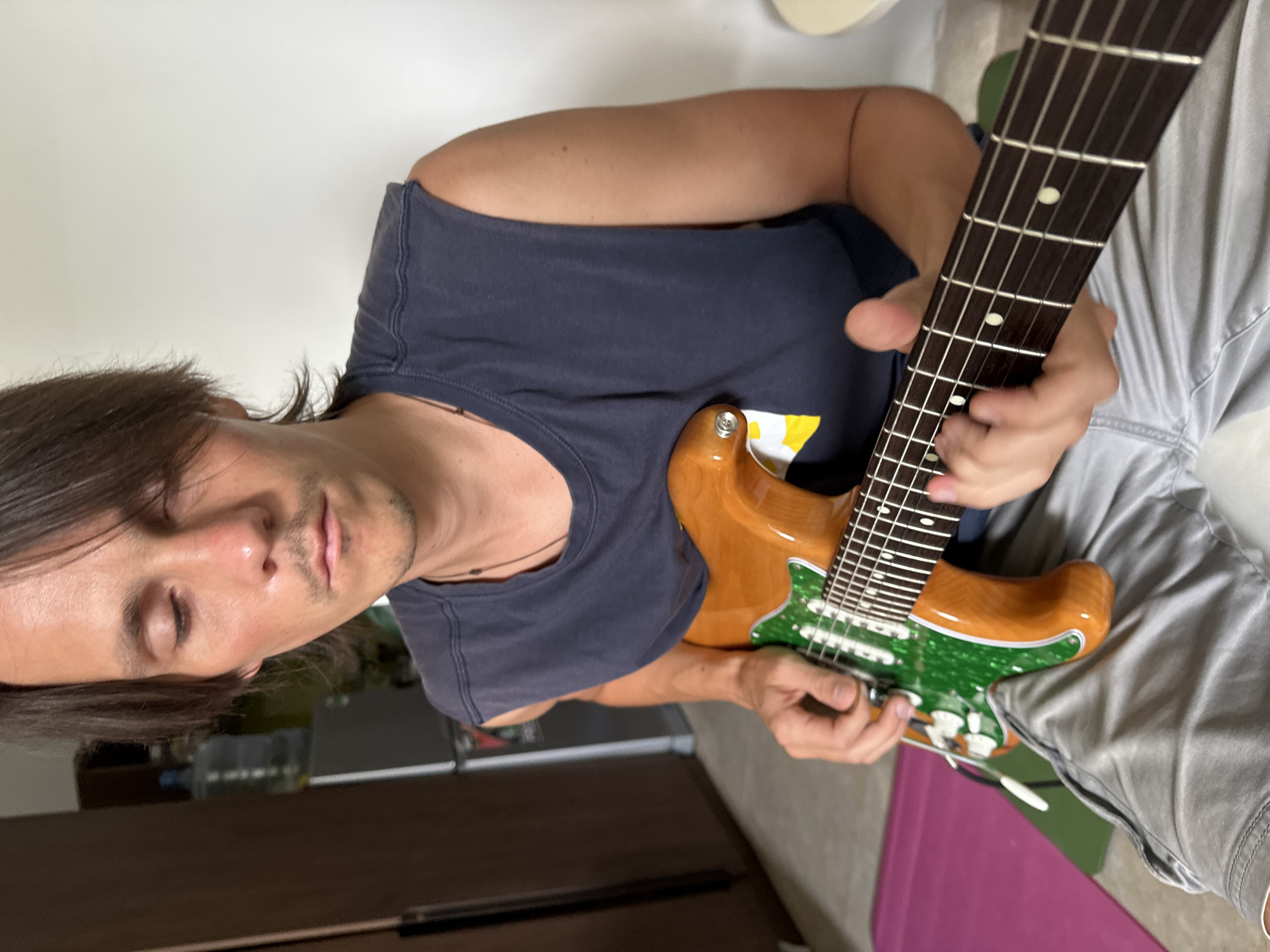 A medium shot shows a man with shoulder-length brown hair and closed eyes playing an electric guitar indoors. He is wearing a dark blue sleeveless shirt and light gray shorts. The guitar has a light brown body and a green pickguard with white pickups. His hands are on the fretboard and near the strings. In the background, a dark brown cabinet or refrigerator is visible to the left, and a white wall is behind the man. A pink mat and a green object are on the floor to the left of the man. The scene appears to be a residential setting in Da Nang, Vietnam.