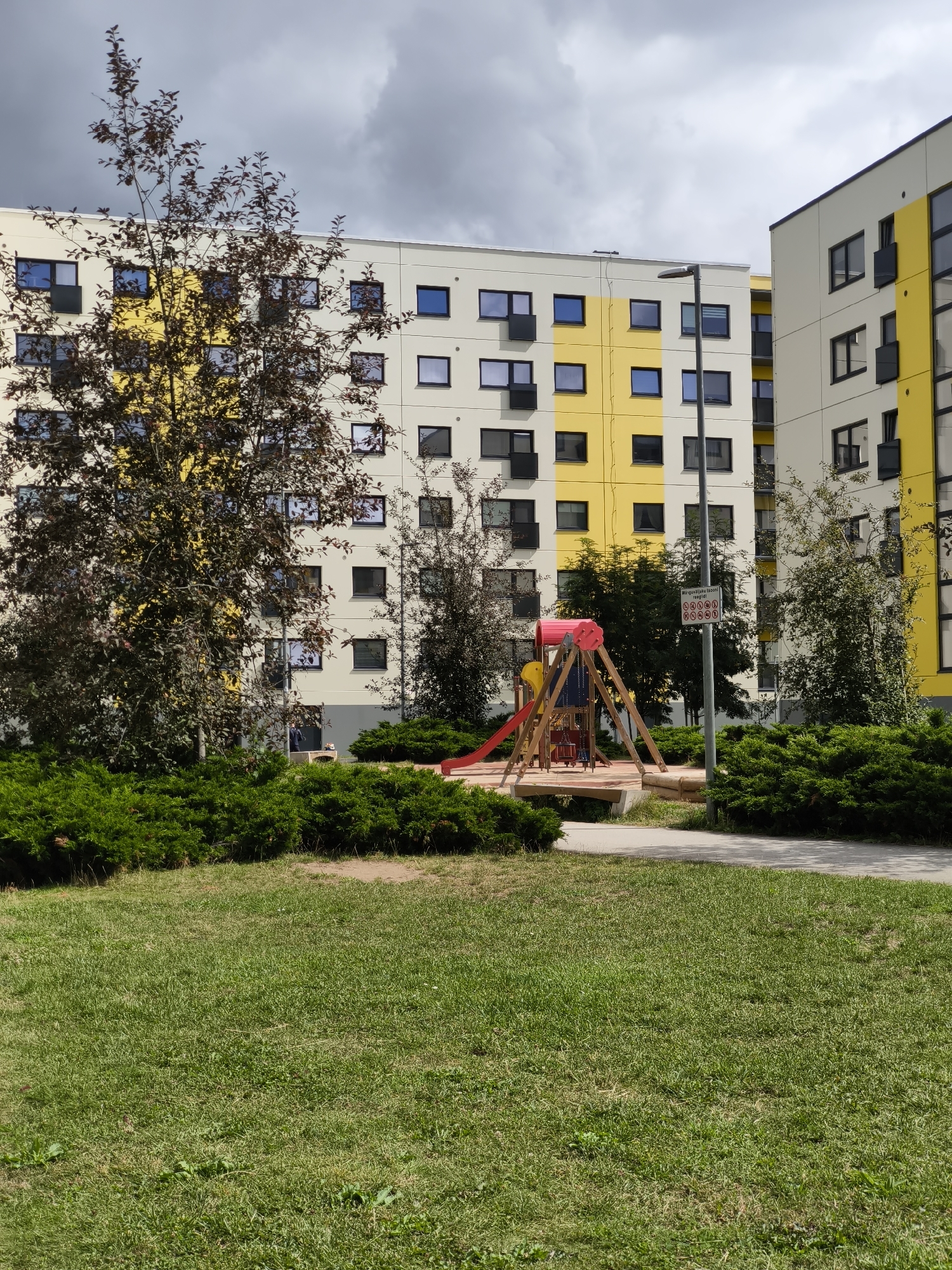 The image depicts a residential area in Tallinn, Estonia, featuring a modern apartment complex with a distinctive yellow and white facade. In the foreground, a well-maintained lawn borders a paved walkway. A children's playground, complete with a slide and swing set, is situated amidst dense green bushes. A solitary street lamp stands near the playground, and a sign with various prohibition symbols is attached to it. The architecture of the buildings suggests recent construction, with numerous windows dotting the facades. The sky is overcast, with dark clouds suggesting imminent rain.