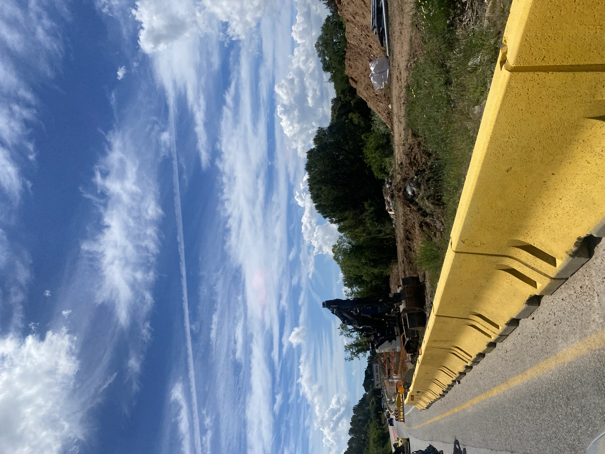 The image depicts a scene along a road in Caldes de Montbui, Spain. A yellow safety barrier runs along the right side of the road. Beyond the barrier, there is a dirt bank covered with grass and some trees. Heavy machinery, including a crane, is visible behind the barrier. Further down the road, a sign points the way to Can Valls. The sky is blue with streaks of white clouds.
