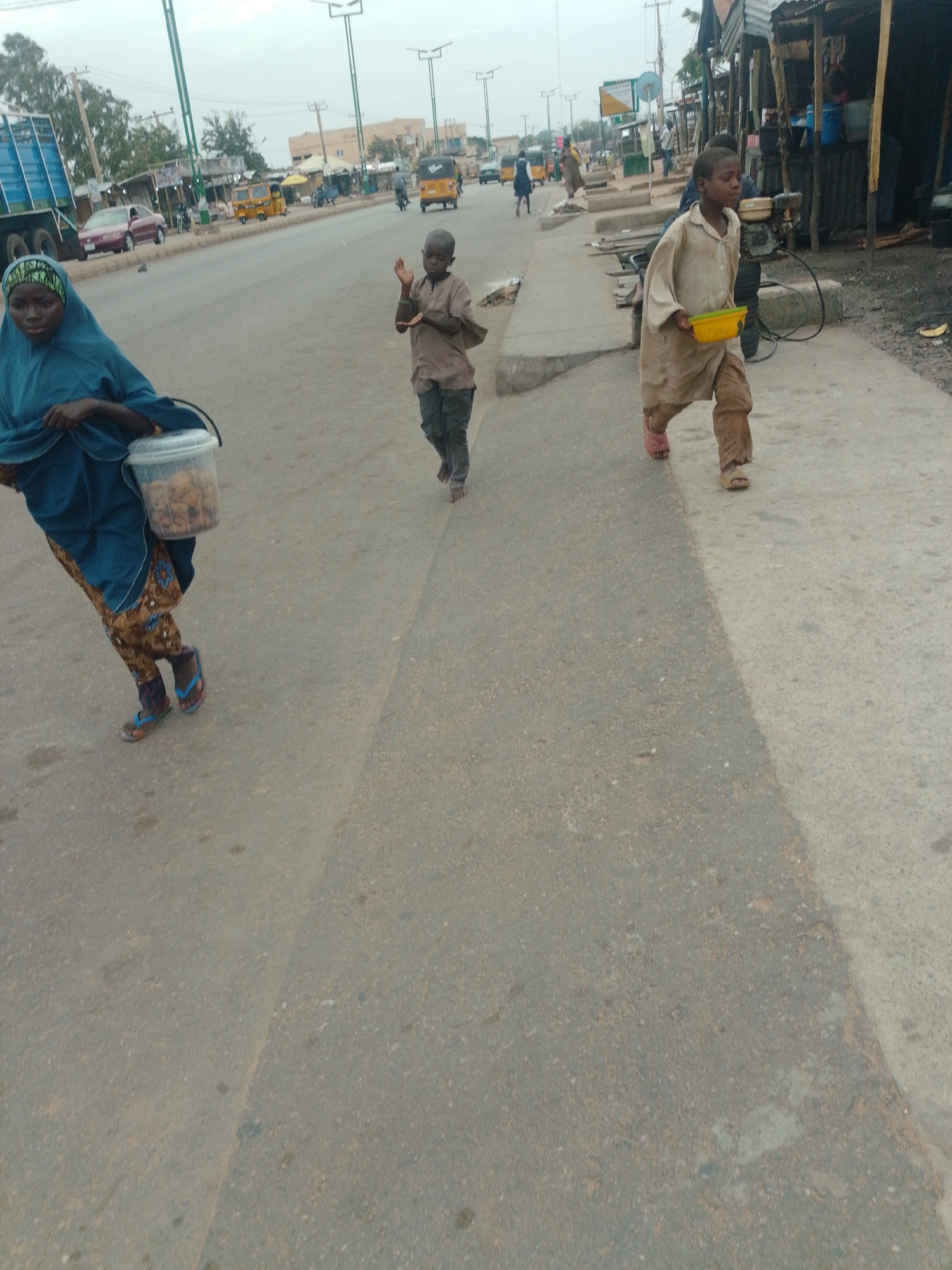 In Gombe, Nigeria, a group of people are seen walking along a paved road. On the left, a woman in a blue hijab and patterned pants carries a clear plastic bucket filled with goods. To her right, a young boy in a brown shirt and dark pants walks ahead, his hands raised as if in greeting. Further ahead and to the right, another boy in a beige, oversized shirt and brown pants walks with a yellow bowl in his hand. The street appears to be lined with market stalls and various vehicles, including a yellow and white taxi, a red car, and a blue truck, suggesting a busy commercial area. The sky is overcast.