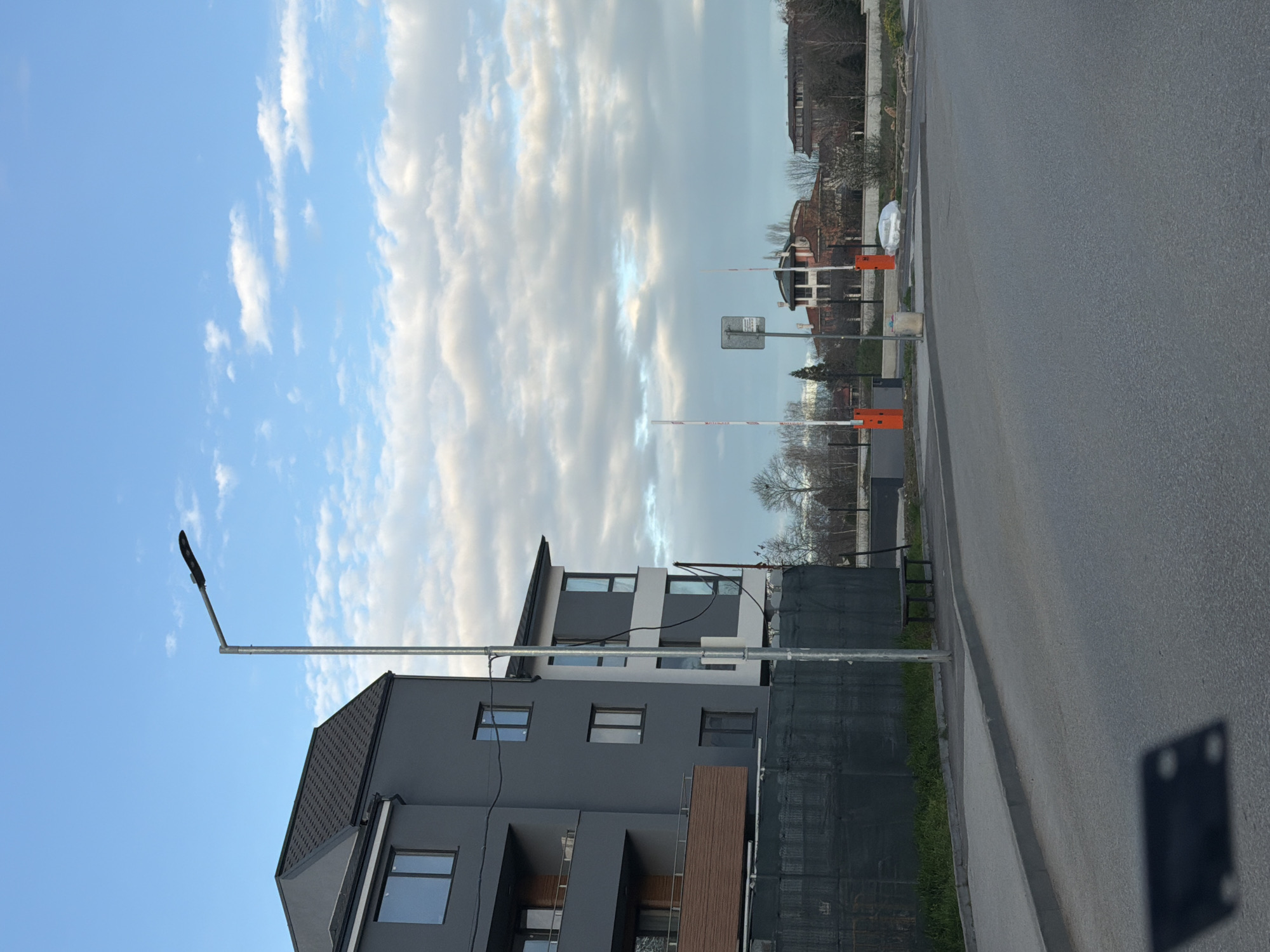 The image shows a street scene in Plovdiv, Bulgaria. A modern, multi-story apartment building with gray exterior and dark roof is on the left. A street lamp extends over the sidewalk in front of the building. A gray asphalt road occupies the right side of the image. In the background, across the road, are barriers, a traffic sign, and indistinct buildings obscured by trees. The sky is partly cloudy.