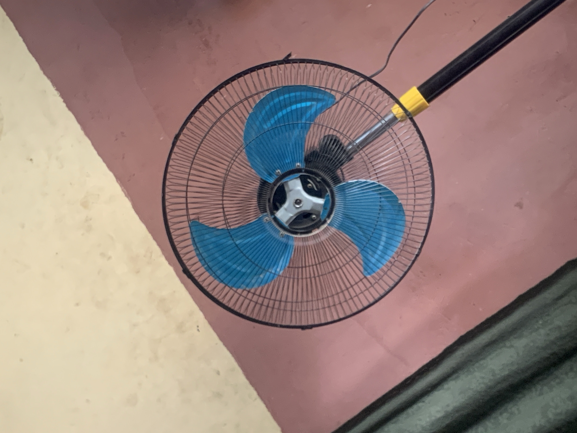 An overhead shot shows a black oscillating fan with bright blue blades and a silver and black center. The fan is positioned on a textured, pinkish-red floor. To the left of the fan, a dark green curtain is visible hanging, creating a diagonal line across the upper left portion of the image. The rest of the floor is a lighter, yellowish-beige color. A black power cord extends from the fan base. The image is taken from an elevated perspective, looking down on the fan and the surrounding floor and curtain. This scene is likely found within a residential or commercial space in Jauro Yinu, Nigeria, an area known for its warm climate, making such fans a common household appliance.