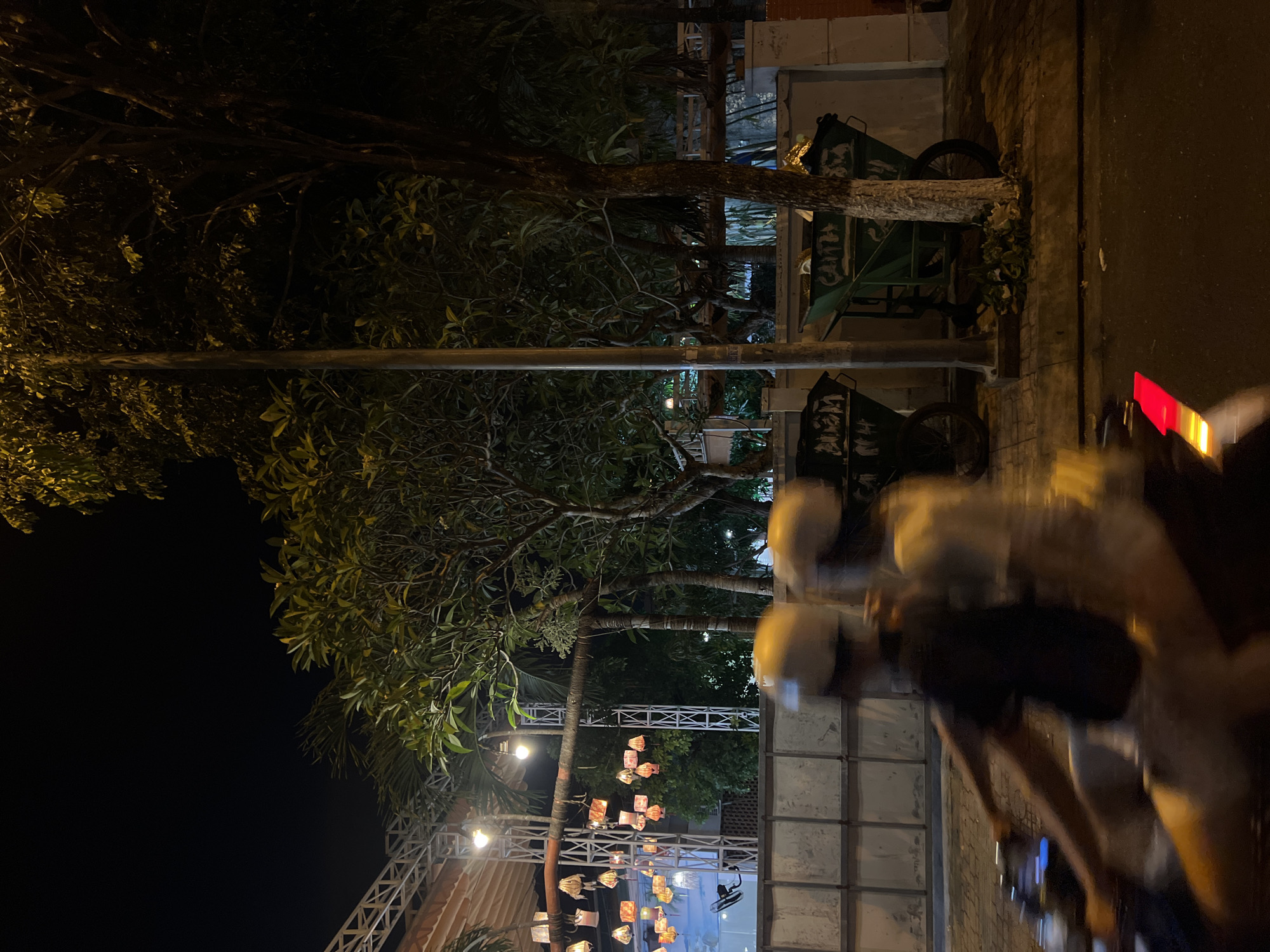 The image depicts an outdoor scene in Nha Trang Ward, Vietnam, at night. The foreground shows pedestrians, likely on a street or sidewalk, with a blurred effect suggesting movement. Next to them is the side of a building, with visible wall textures. Trees with leaves are present, with some lights or lanterns hanging from the branches of trees or nearby structures. Additionally, there are green carts or vehicles. The image is rotated.