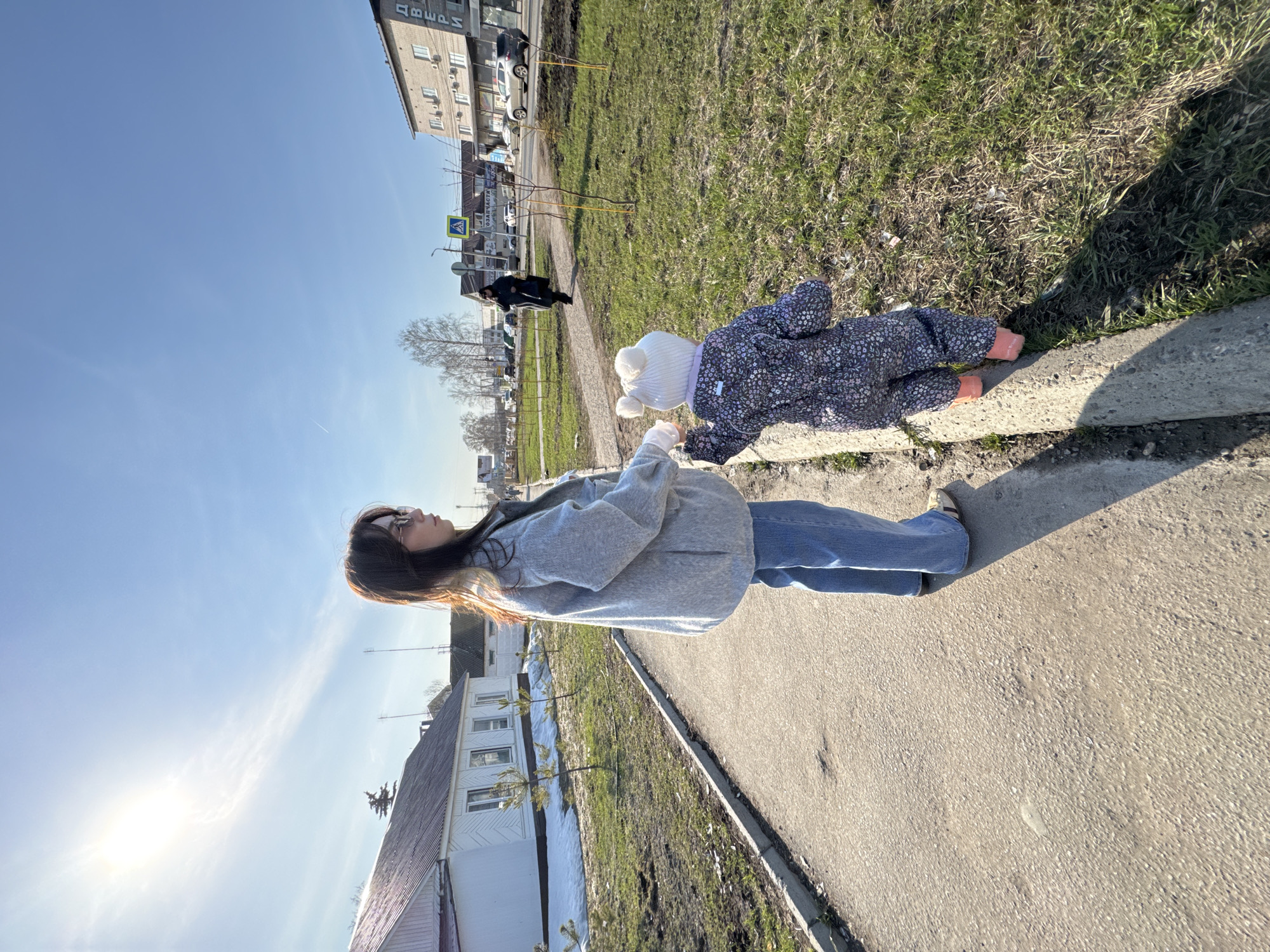 The image shows a young woman and a toddler walking along a sidewalk in Chaikovsky, Russia. The woman is wearing a light gray hooded sweatshirt, blue jeans, and sneakers. She has long dark hair and is wearing glasses. The toddler is wearing a dark blue floral-patterned snowsuit, a white knit hat with pom-poms, and orange booties. The toddler is leaning over the edge of the sidewalk, looking at the grass. The woman is holding the toddler's hand. The sidewalk is next to a grassy area with a concrete curb. In the background, there is a street with buildings, cars, and people. The sky is blue with some clouds. The sun is shining.