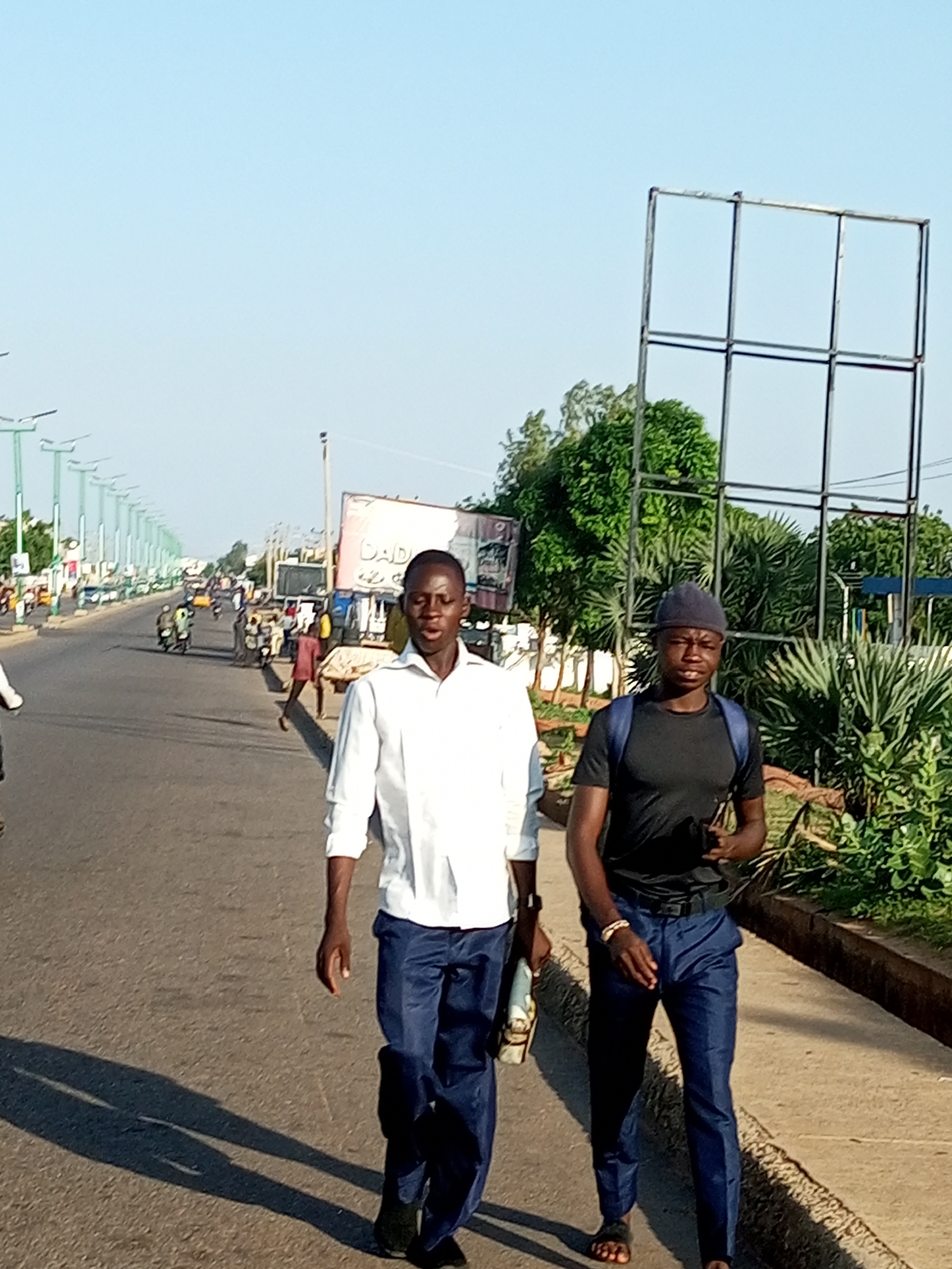 Two young Black males are walking on a paved road in Tempure, Nigeria. The male on the left is wearing a white long-sleeved shirt and dark blue pants, and is carrying a white bag. The male on the right is wearing a black t-shirt, dark blue pants, and a black beanie, and has a blue backpack. They are walking on the right side of the road, on a sidewalk. Behind them, on the left side of the road, there is a row of streetlights and trees, as well as other people and vehicles, including motorcycles and cars. On the right side of the road, there are lush green trees and plants. The sky is clear and blue. There is a large metal structure in the background, which may be a billboard frame. The scene appears to be a street in a bustling urban area.