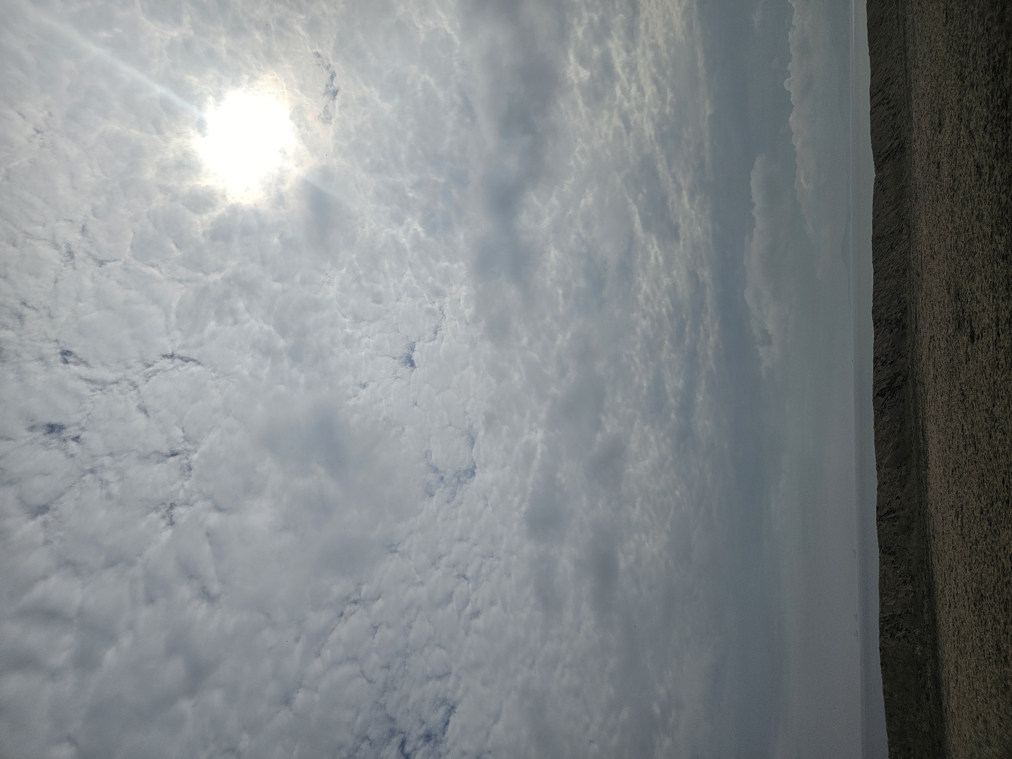 The image depicts a cloudy sky with the sun shining through, potentially in Burgas, Bulgaria. A strip of land or coastline is visible on the right side of the frame. The sky is predominantly filled with a blanket of small, rounded clouds, creating a textured, overcast appearance. The sun is bright and partially obscured by the clouds, casting a slight glare. The ground or coastline appears rough and textured.