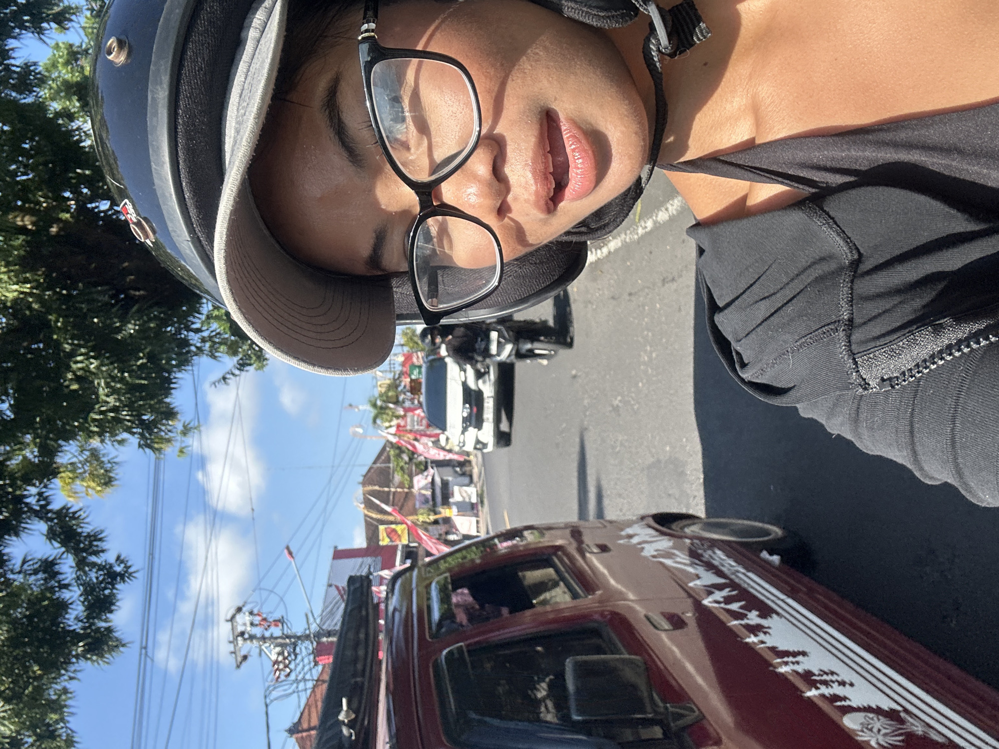 In Sukawati, Indonesia, a woman wearing a helmet and glasses is riding a motorbike on a street. She is in the foreground, facing right and looking at the camera. Behind her, a red car is visible on the left side of the image, with a white mountain and palm tree decal on its side. In the background, several buildings and a white car are visible on the street, which is lined with red and white flags. The sky is blue with some clouds, and trees are present in the upper part of the image. The scene appears to be a street in a bustling area, possibly a market or commercial district.