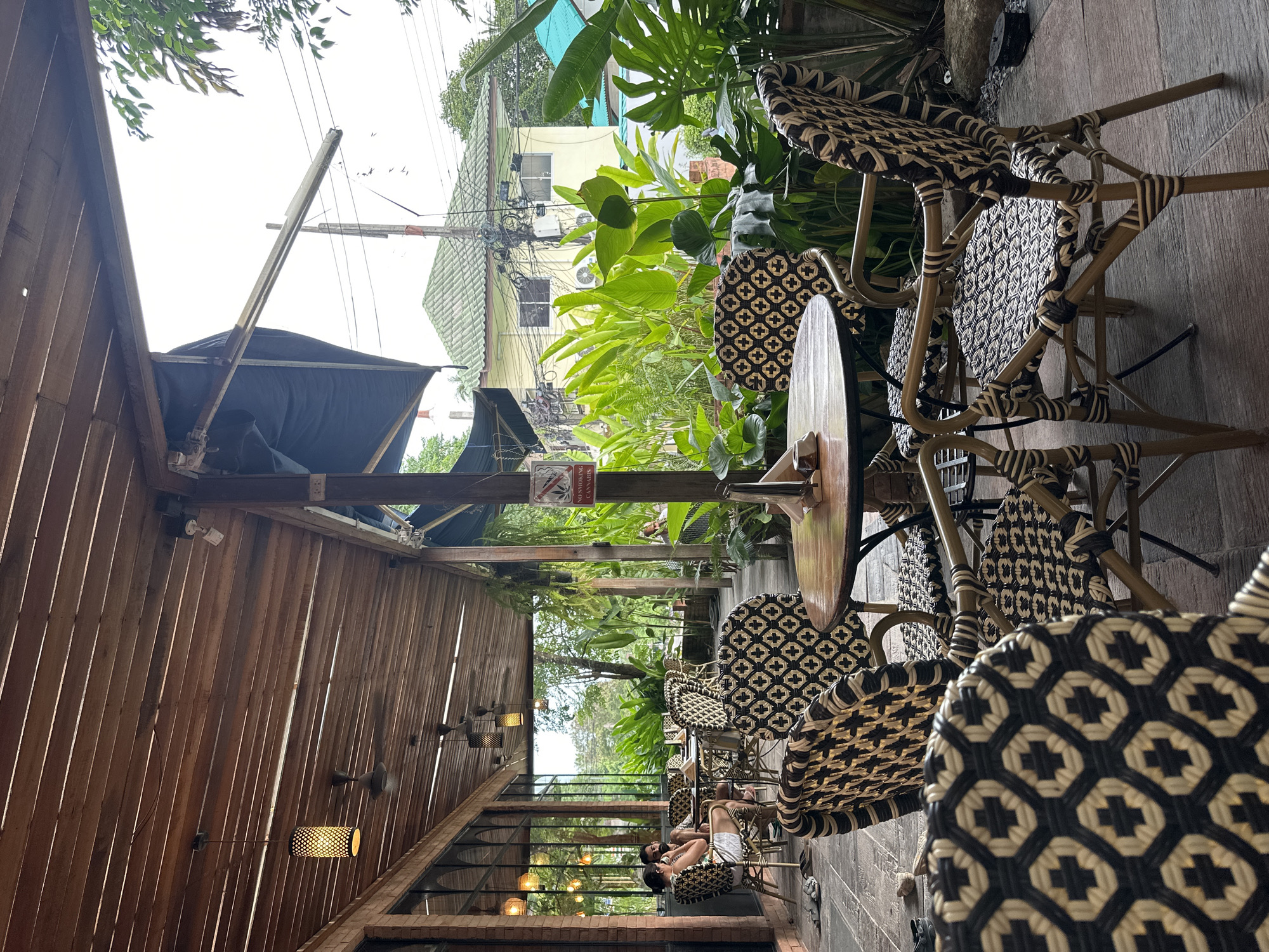 This image depicts an outdoor seating area of a restaurant or cafe in Koh Phangan, Thailand. The scene is characterized by a covered patio with a wooden slatted ceiling, illuminated by pendant lights and ceiling fans. Woven rattan chairs with black and cream patterned cushions surround wooden tables. In the background, lush green tropical foliage is visible, with a glimpse of a building with a green tiled roof and yellow walls. A "No Smoking Cannabis" sign is posted on a wooden pole. Two individuals are seated at a table in the middle ground, engaged in conversation. The flooring is stone tile.
