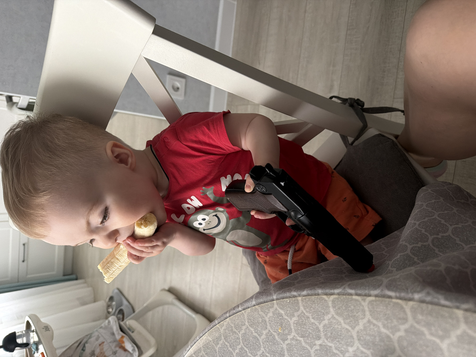 A young child with fair hair is sitting in a highchair, eating a banana with their left hand and holding a black handgun in their right. The child wears a red t-shirt with a cartoon monkey graphic and orange shorts. The highchair has a white frame and a gray patterned seat. The background shows a light-colored wall and furniture. The scene is not specified as being in Votkinsk, Russia.