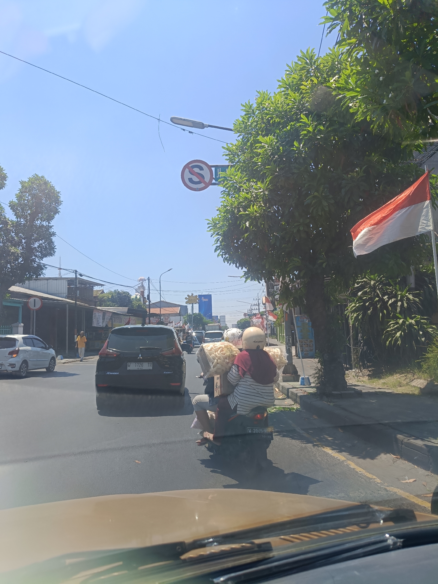 In Sidoarjo, Indonesia, on a clear, sunny day, a street scene unfolds with moderate traffic. The foreground shows the dashboard of a vehicle, suggesting the photo was taken from inside a car. A dark-colored SUV with license plate "W 1726 TR" is in the immediate vicinity on the asphalt road, followed by a motorcycle carrying two passengers and a substantial load of puffed snacks. The rider wears a helmet and a hijab, while the passenger wears a striped shirt and also has a helmet. A large tree with dense green foliage dominates the right side of the image, partially obscuring the sky and a red and white Indonesian flag attached to a pole. On the left side of the road, a white hatchback is parked, and a man in a yellow shirt walks along the sidewalk. Buildings line the street, some with awnings and signs, indicating commercial activity. Traffic signs, including a "no U-turn" symbol, are visible along the road. The environment appears to be a bustling urban or semi-urban area.