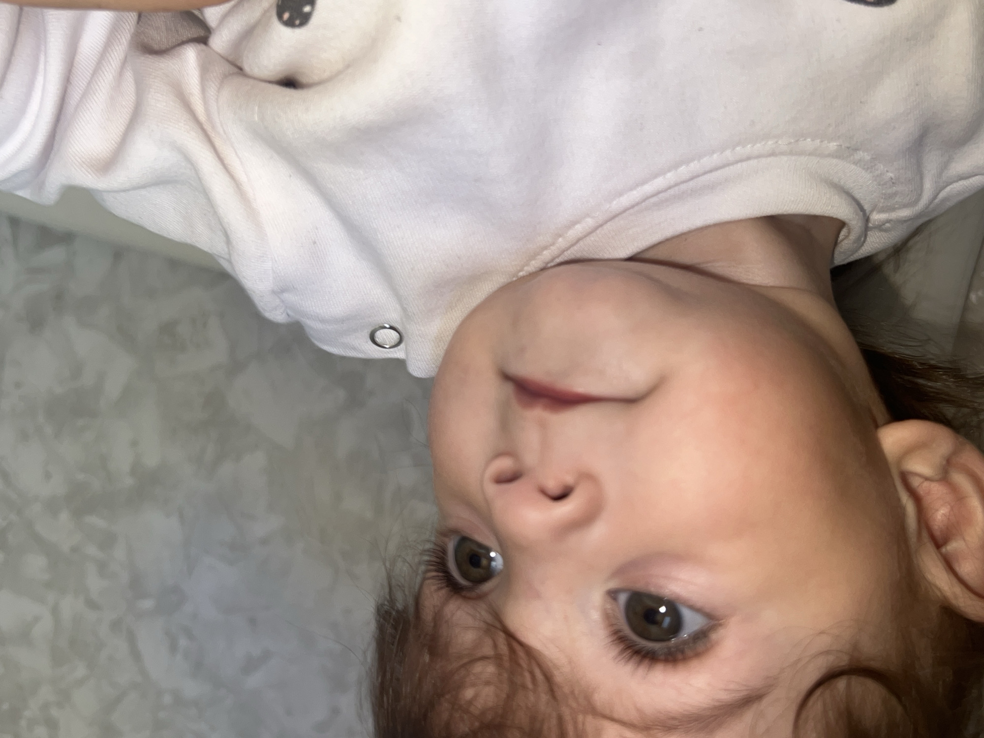 A close-up shot shows the face of a young child with brown eyes and hair, wearing a white top with a button detail. The child's head is tilted, with the face and eyes appearing centered in the frame. The background consists of a light-colored, textured surface. No information is discernible in the image regarding Elena, Bulgaria.
