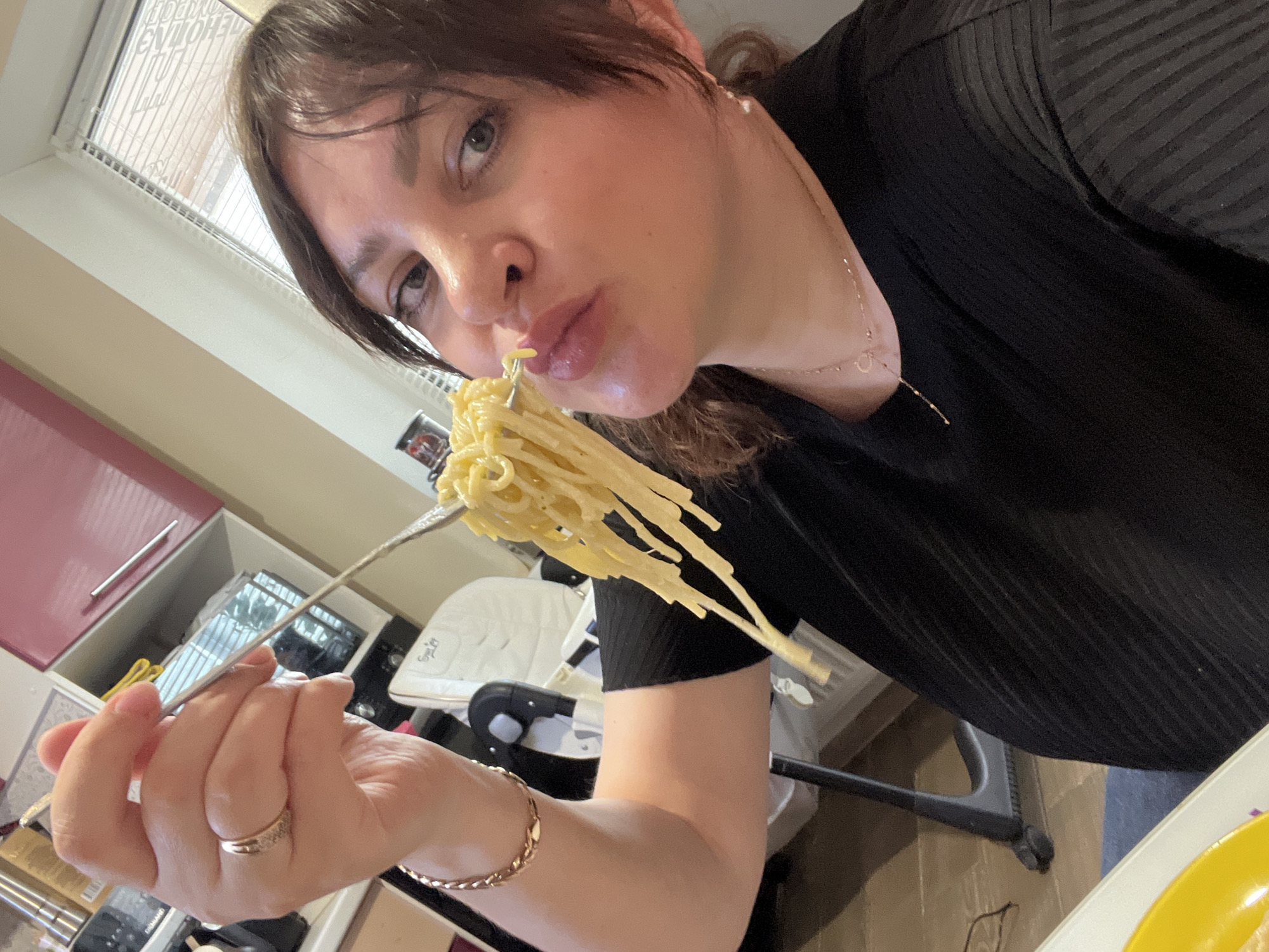 A woman with dark hair and fair skin holds a fork laden with pasta near her mouth, positioned indoors. She is wearing a dark-colored top and jewelry including a ring and a bracelet. A child's high chair is visible in the background, as well as cabinets, a window, and various household items. The location is identified as Noviy, Russia, but no information to confirm this from the picture.