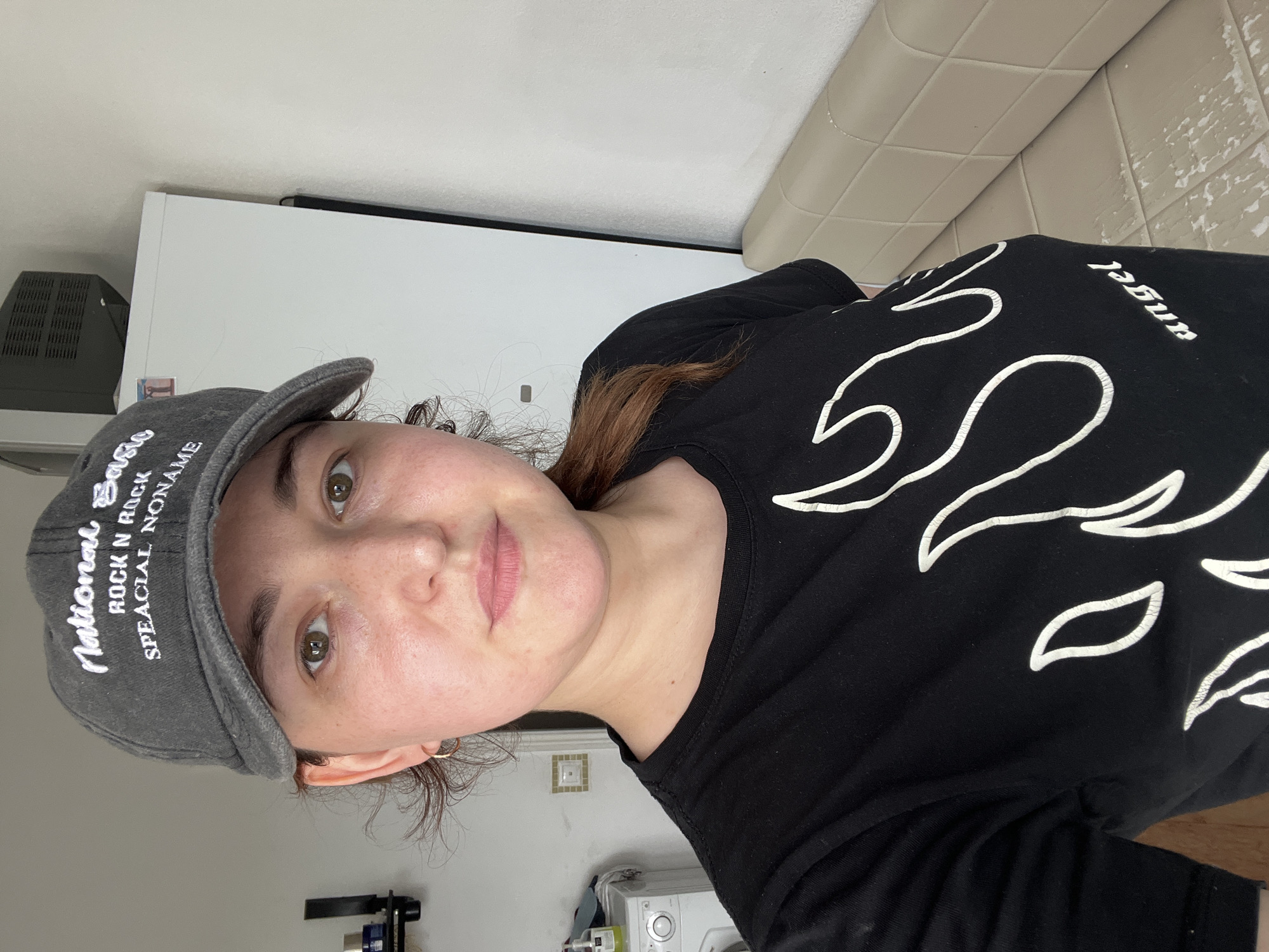 A young woman is taking a selfie in what appears to be a residential interior. She is wearing a grey baseball cap with white lettering that reads "National Basic Rock N Rock Special Noname" and a black t-shirt with a white flame design and the word "angel" on it. She has light brown hair, brown eyes, and is wearing a gold hoop earring. The background includes a white appliance, possibly a washing machine, to her left, a white closet or refrigerator to her right, and a cream-colored tufted bench. The lighting suggests it is daytime. There is no indication that this image was taken in Chelyabinsk, Russia.