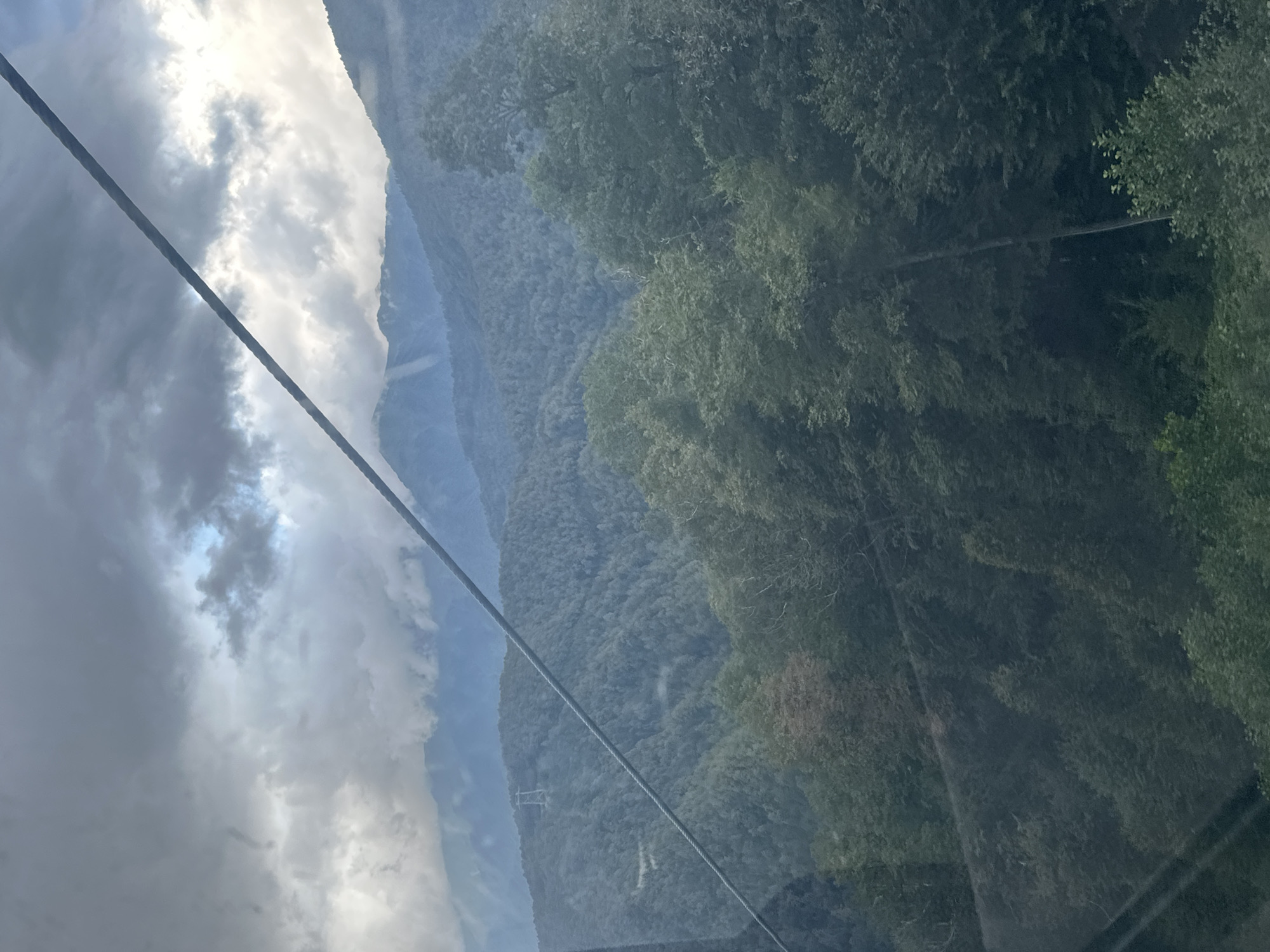 The image captures a scenic view of the mountainous landscape surrounding Estosadok, Russia. A thick, dark cable stretches diagonally across the frame from the upper left to the lower right, indicating the perspective is likely from a cable car or similar transportation. The mountains are densely covered with lush green trees, creating a textured, undulating terrain. In the mid-ground, a solitary power line pylon stands on one of the slopes, a man-made structure amidst the natural beauty. The sky above is dramatic, filled with heavy, dark clouds that partially obscure the sun, allowing shafts of light to break through in places, illuminating parts of the distant mountain peaks with a soft, ethereal glow. The overall impression is one of a vast, somewhat moody, natural environment.