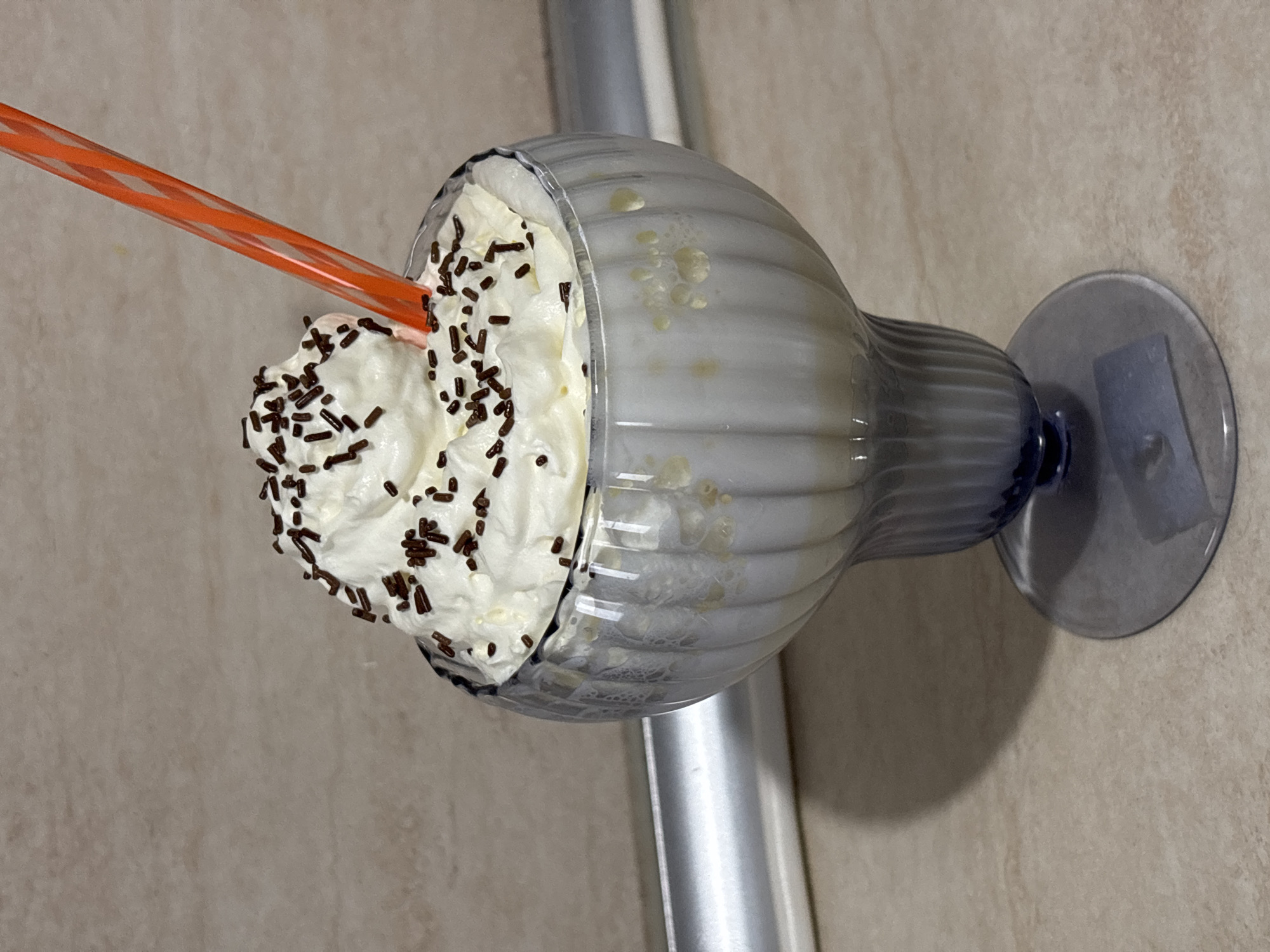 A dessert drink is displayed in a clear glass with a blue base, likely a milkshake, topped with a generous amount of whipped cream and chocolate sprinkles. An orange and white striped straw is inserted into the whipped cream. The glass rests on a surface with a tan-colored background, potentially a counter or tabletop. A metallic, cylindrical object is positioned behind the glass, adding a subtle element to the scene. The city and location of Unknown, Unknown cannot be verified in the scene.
