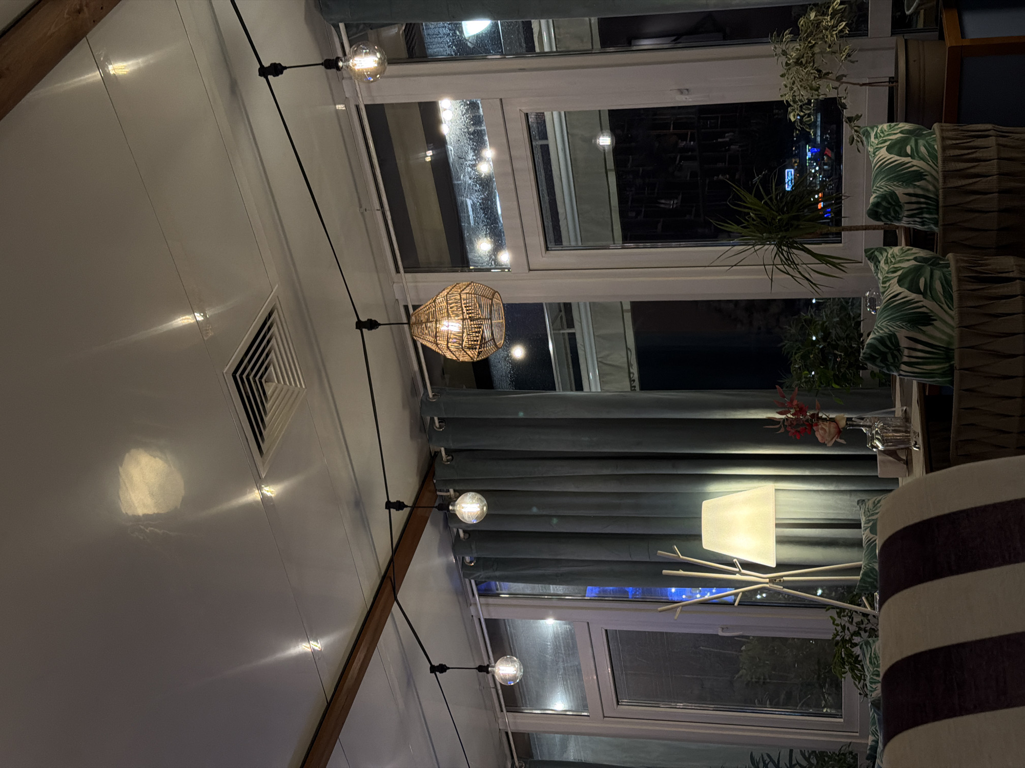The image displays an interior view of a commercial establishment, likely a restaurant or cafe, located in Varna, Bulgaria, during evening or nighttime hours. The white ceiling features a central air vent and visible reflections from artificial light sources. A dark overhead cable suspends multiple exposed spherical light bulbs and one woven rattan pendant lamp. A horizontal wooden beam is visible in the upper left quadrant. Large, white-framed windows, partially obscured by teal-colored curtains, dominate the midground. The exterior view through the windows is dark, showing reflections of interior lights and distant, blurred urban illumination. Interior decor includes a white floor lamp with a conical shade providing localized light, positioned next to a white, stylized, multi-pronged coat rack. Seating in the foreground consists of a bench with maroon and cream striped cushions. Additional cushions featuring a green palm-leaf pattern rest on wicker-textured bases. Potted plants, including a tall Dracaena and a variegated Ficus, are strategically placed near the windows. A small glass vase containing red and pink flowers is visible on a table surface. No individuals are present in the scene.