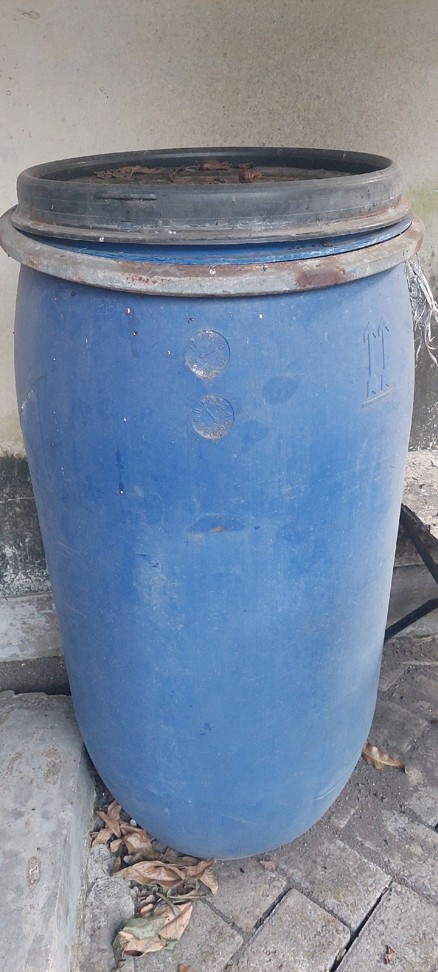 The image shows a blue plastic barrel with a black lid, situated outdoors. The barrel has some markings on its side, including two circular stamps and an upward-pointing arrow symbol. The lid appears to be rusty around the rim. The barrel is placed on a ground covered with interlocking paving stones and some dry leaves have fallen around its base. In the background, a textured concrete wall is visible. The scene is likely located in Lumajang, Indonesia, given the context of the request. There are no people or animals visible in the image. The lighting suggests it is daytime.