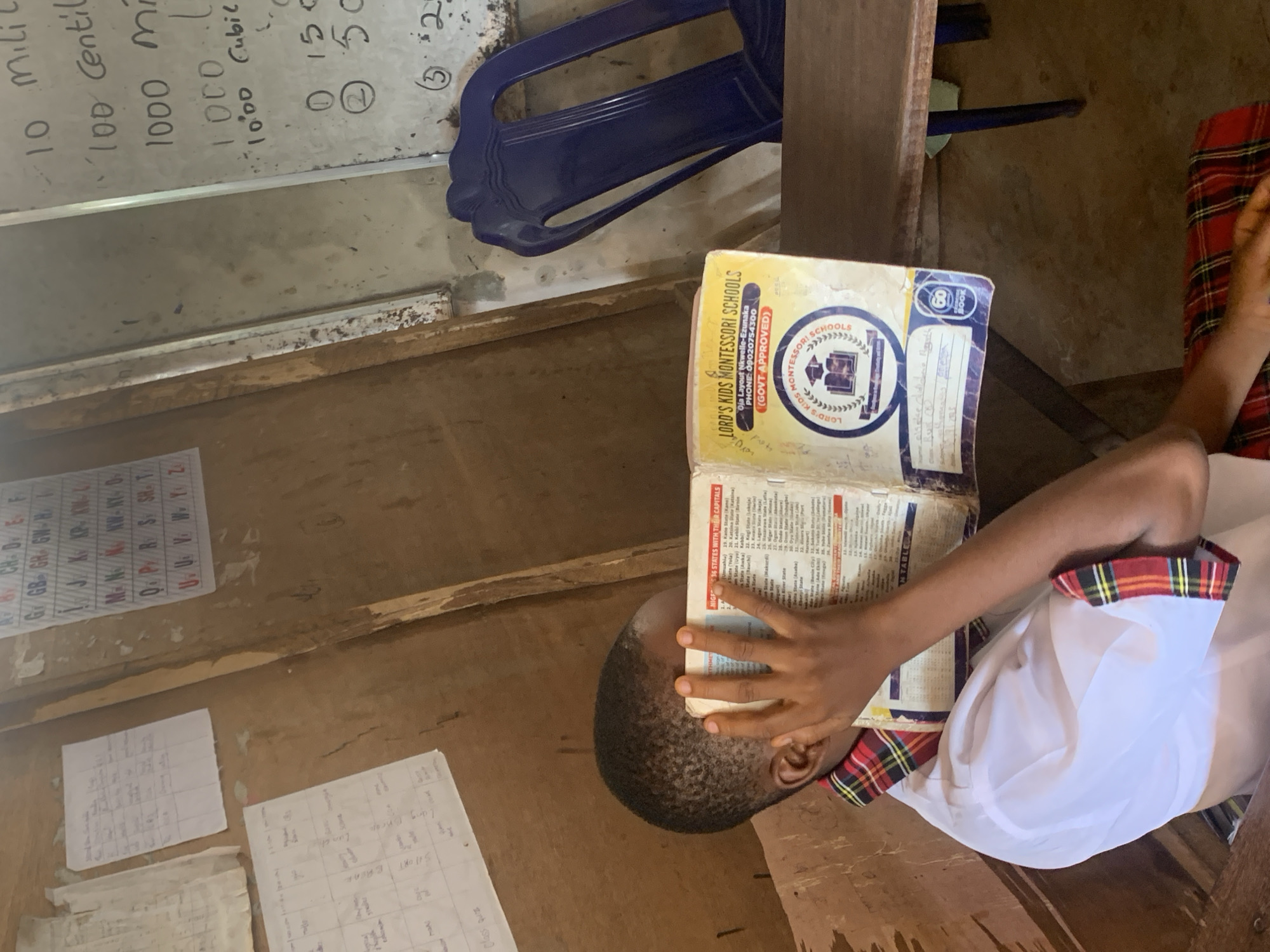 A child sits at a desk in what appears to be a classroom. The child is reading a book with "NIGERIA's 36 STATES WITH THEIR CAPITALS" printed on one of the pages. The book has the "LORD'S KIDS MONTESSORI SCHOOLS" logo on the cover. In the background, a whiteboard with math equations written on it can be seen. There are sheets of paper with handwritten notes attached to the wall, including an alphabet chart. A blue plastic chair is partially visible above the book. The classroom environment suggests a school setting in Nigeria. There is no further information provided about a specific location in the city of Ukwu-Abwa, Nigeria.