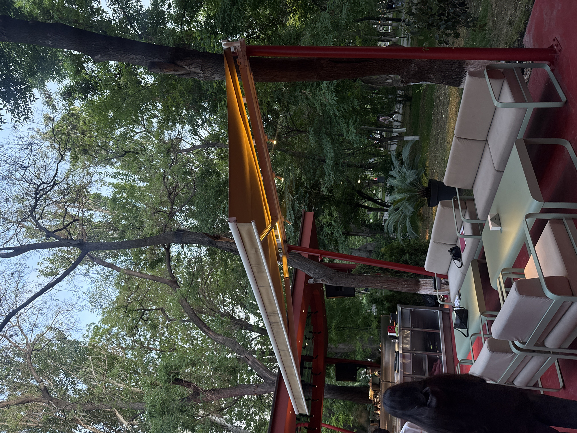 The image shows an outdoor seating area, possibly a café or restaurant, located in Yerevan, Armenia. The area is covered by a brown awning with a white underside, supported by red metal beams. The seating area includes light-colored sofas and chairs with green metal frames, arranged around matching tables. The floor is a red surface. Trees are visible overhead, providing shade to the area, and a bar is set up in the background. Individuals can be seen at the bottom of the frame.