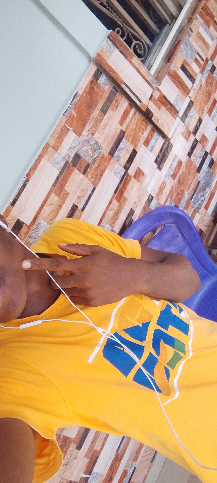 A person wearing a yellow t-shirt with a graphic design is lying down on a blue plastic chair. The person's right hand is raised, with the index finger pointing upwards. White earbuds are in the person's ears and draped around their neck and chest. The background consists of a wall tiled with rectangular pieces in various shades of brown, beige, and gray. A dark decorative metal railing is visible at the top of the image. The scene appears to be outdoors, possibly on a balcony or patio.