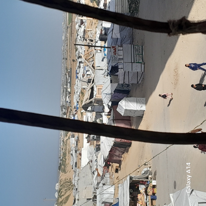 An encampment of makeshift dwellings stretches across a barren landscape under a clear sky. Structures of white and gray tarpaulin dominate the view, interspersed with wooden frameworks and piles of materials. People are walking on a sandy path alongside the encampment. The scene is framed by dark, out-of-focus wooden structures.