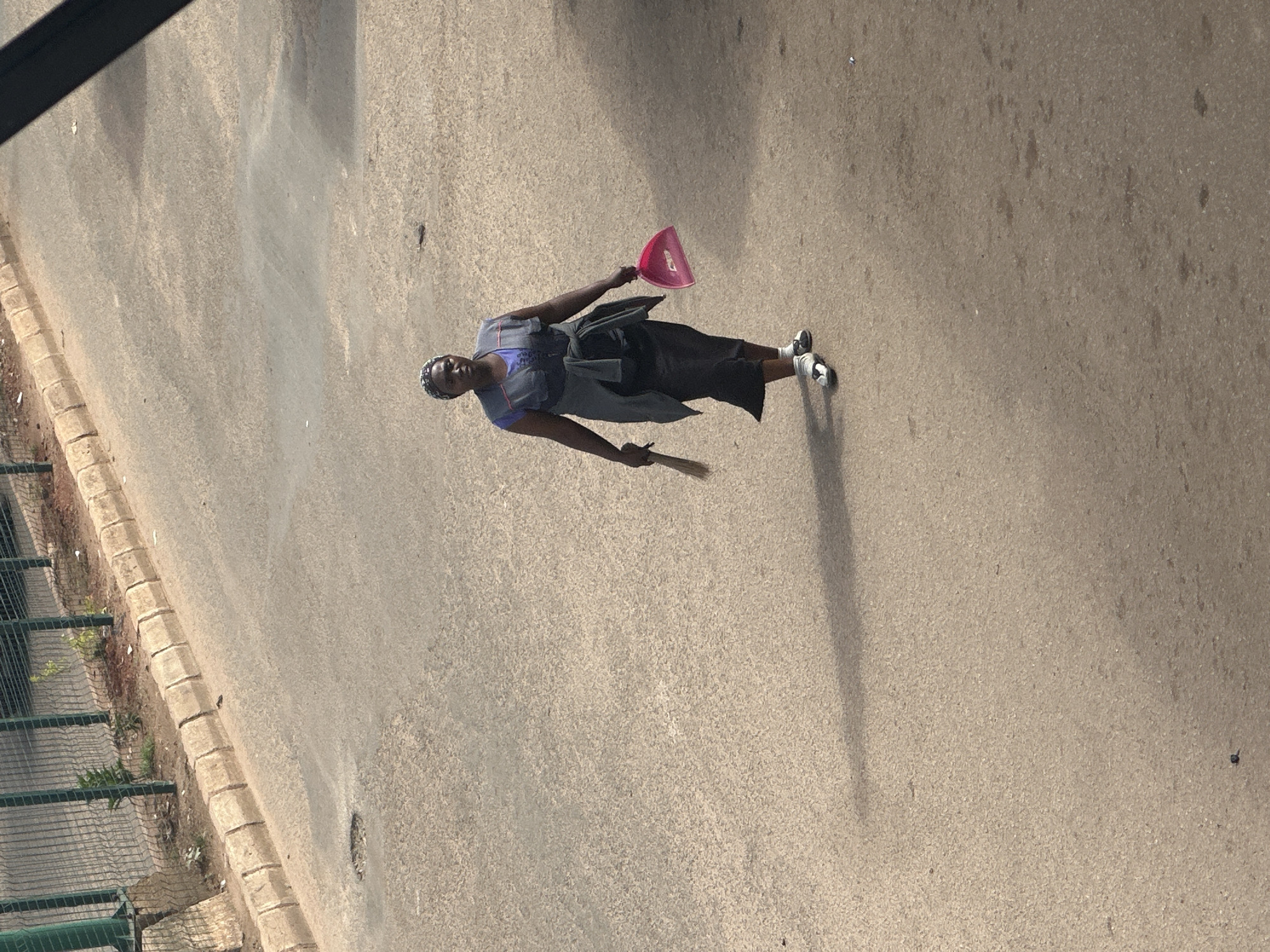 A woman walks across a paved street in Bukuru, Nigeria, holding a broom and a pink dustpan. She is wearing a blue shirt, a grey vest, and a black skirt, with a grey sweater tied around her waist. The street is somewhat worn, with a small pothole visible to the left of the frame. A green metal fence runs along the side of the road. The scene appears to be captured from an elevated vantage point, casting a long shadow of the woman onto the pavement.