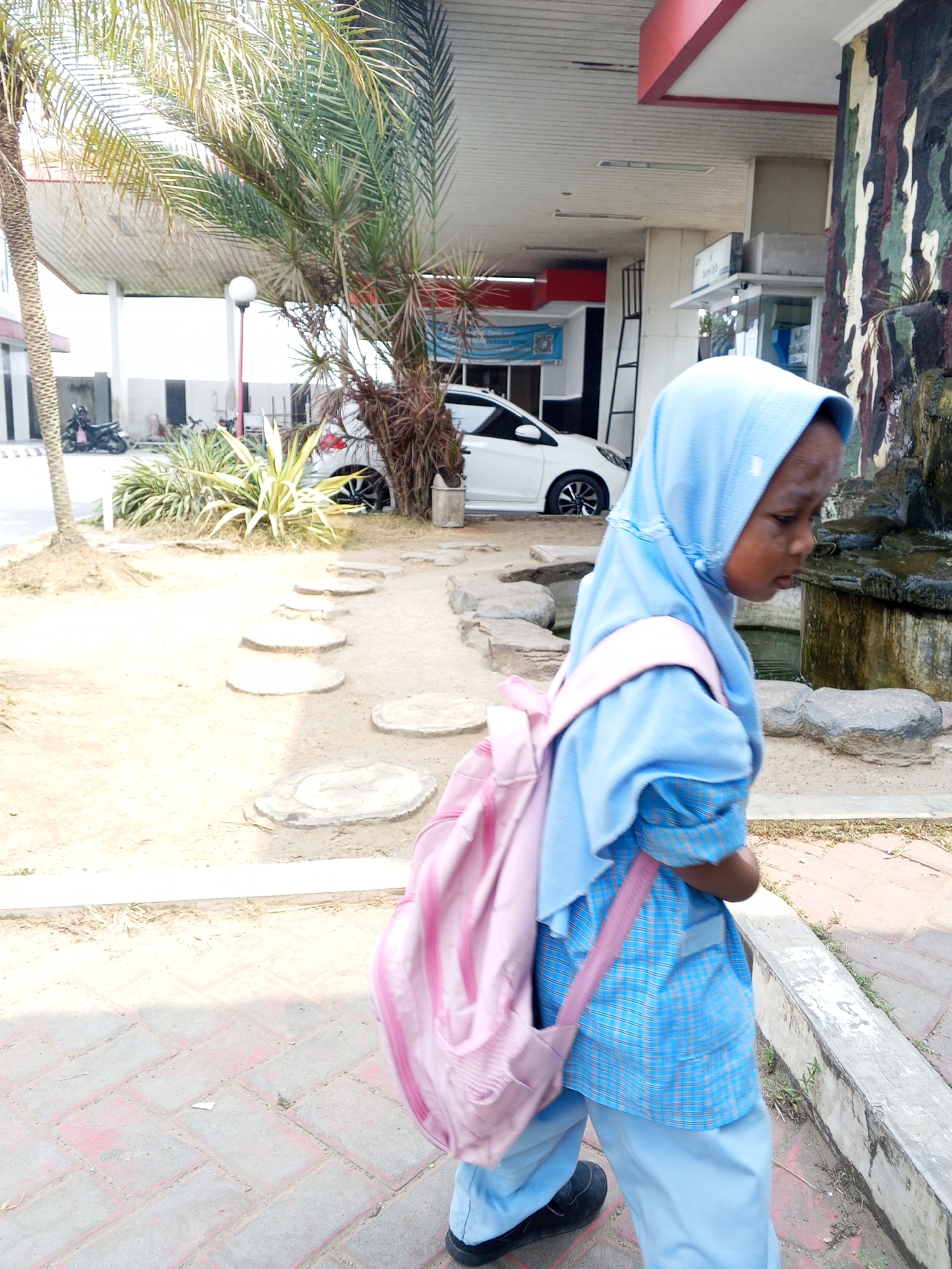 In Ciruas, Indonesia, a young child wearing a blue hijab and a blue plaid shirt with light blue pants walks to the right, carrying a pink backpack. The child is walking on a paved walkway made of red bricks. To the left of the walkway is a sandy area with stepping stones. In the background, a white car is parked, and a palm tree stands tall. The scene appears to be outside a building with a red and white awning.