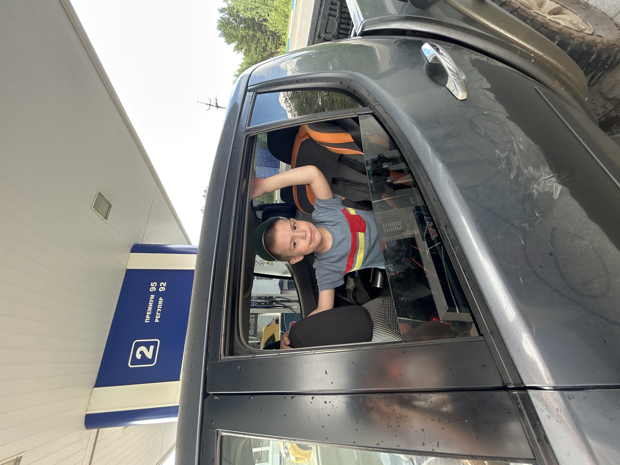 A young boy wearing a green baseball cap and a grey t-shirt with horizontal red, yellow, and blue stripes is visible through the open window of a dark grey pickup truck. He is leaning against the car door, with his left hand resting on the window frame. The boy appears to be looking towards the camera with a slight smile. The truck is parked at a gas station, indicated by the blue and white sign with the number "2" and text in Russian that reads "ПРЕМИУМ 95" (PREMIUM 95) and "РЕГУЛЯР 92" (REGULAR 92). The background shows a gas pump and other vehicles, as well as trees and a utility pole, suggesting an outdoor environment. The sky is overcast, and there are water droplets on the truck's windows. The overall scene captures a moment at a gas station in the city of Chaikovsky, Russia.