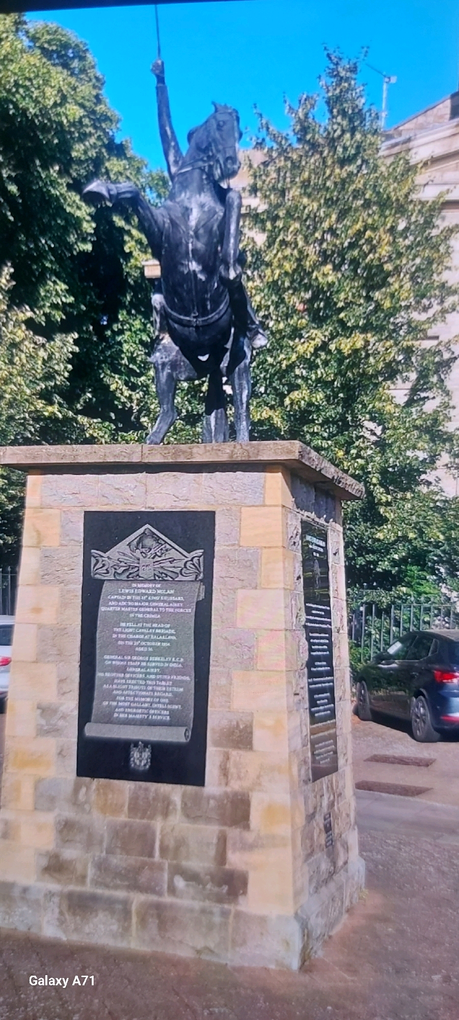 This is a vertical, slightly blurry, exterior daytime photograph featuring a bronze equestrian statue atop a stone pedestal. The statue depicts a figure on horseback, with the rider's left arm raised, holding a sword. The horse appears to be rearing on its hind legs. The statue is dark and weathered.
The stone pedestal is constructed from light-colored, rough-hewn blocks, some of which are accented with darker stones. A black plaque with scrolling text and a decorative emblem is affixed to the front of the pedestal. To the right of this plaque, on the side of the pedestal, is another, longer black plaque with text.
Behind the pedestal, lush green trees with dense foliage are visible, with a portion of a building peeking through the branches. A dark-colored car is parked on the right side of the frame, next to a metal fence. The sky is a bright, clear blue.
There is no indication in the image that this scene is located in Moscow, Russia. The text on the plaque is difficult to read clearly due to the image quality and angle, but it appears to be a memorial inscription. The text on the main plaque begins with "IN MEMORY OF LEWIS EDWARD M'CLAN..." and mentions "CAPTAIN IN THE 11th KING'S HUSSARS." The car and the architectural style of the building do not suggest a Russian context.