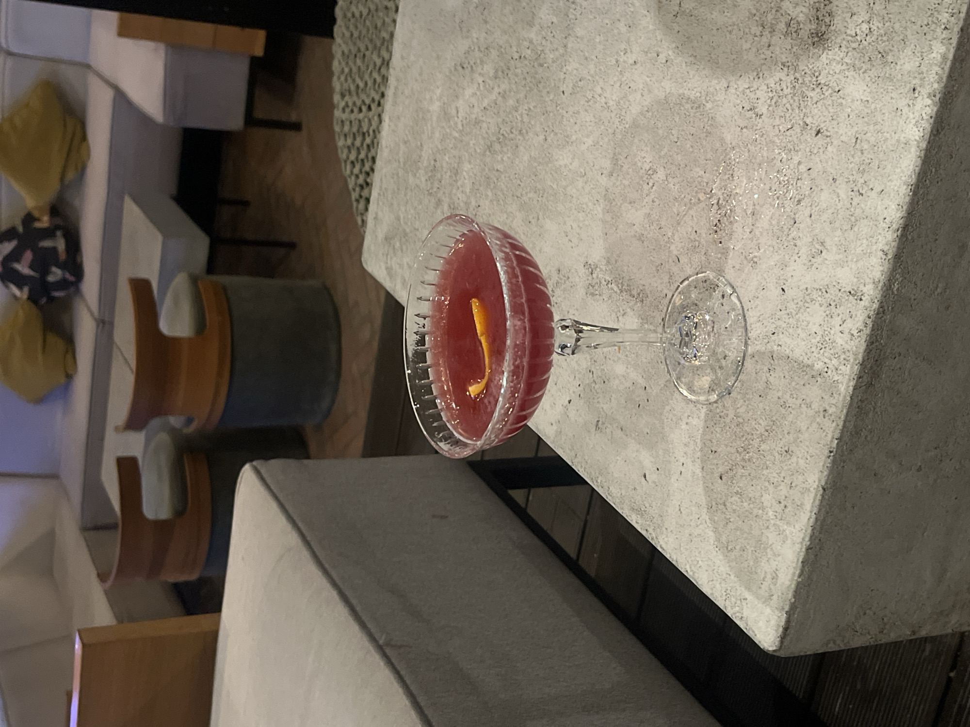 A close-up, eye-level shot captures a vibrant red cocktail in a coupe glass, adorned with an orange peel garnish. The glass rests on a light gray, textured surface, possibly concrete, with a subtle sheen suggesting it may be damp. In the background, soft focus reveals a contemporary lounge area with muted tones. Light gray upholstered seating arrangements are visible, along with low tables and two distinct stools with reddish-brown tops and dark gray cylindrical bases. A wooden partition is present on the left side of the frame. The overall ambiance appears to be that of a modern establishment, possibly a bar or lounge in Burgas, Bulgaria.